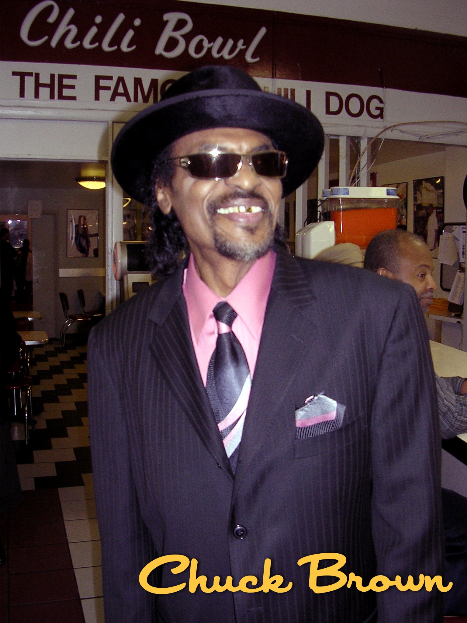 A man dressed in a black pinstripe suit, pink shirt, and black tie, wearing a black hat and sunglasses, smiling with gold teeth, standing inside a restaurant with a sign that reads "Chilli Bowl" in the background.