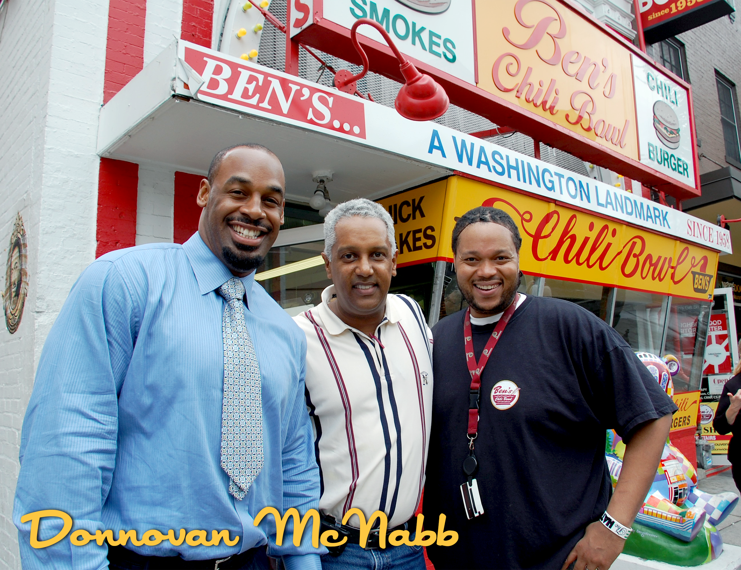 Three men standing in front of Ben's Chili Bowl, smiling at the camera. The storefront has colorful signs and a large banner with the name of the restaurant. The men are dressed casually and appear happy.