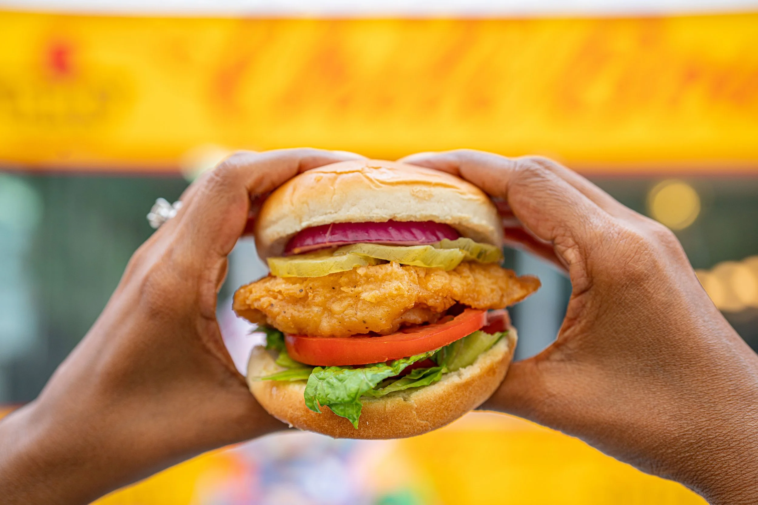 Close-up of a person holding a fried chicken sandwich with pickles, tomato, lettuce, and red onion in a bun.