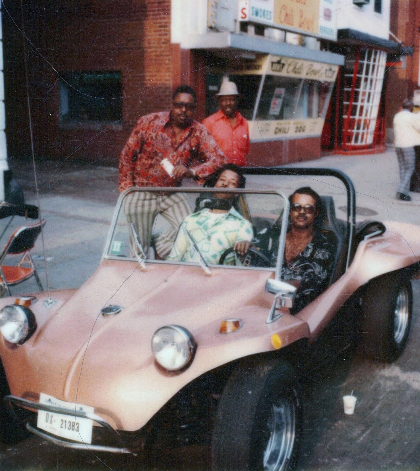 Four people with dark skin and afro hairstyles outside a brick building with a storefront, two men standing and two women sitting in a pink dune buggy with a clear windshield, one woman wearing glasses, in an urban setting.