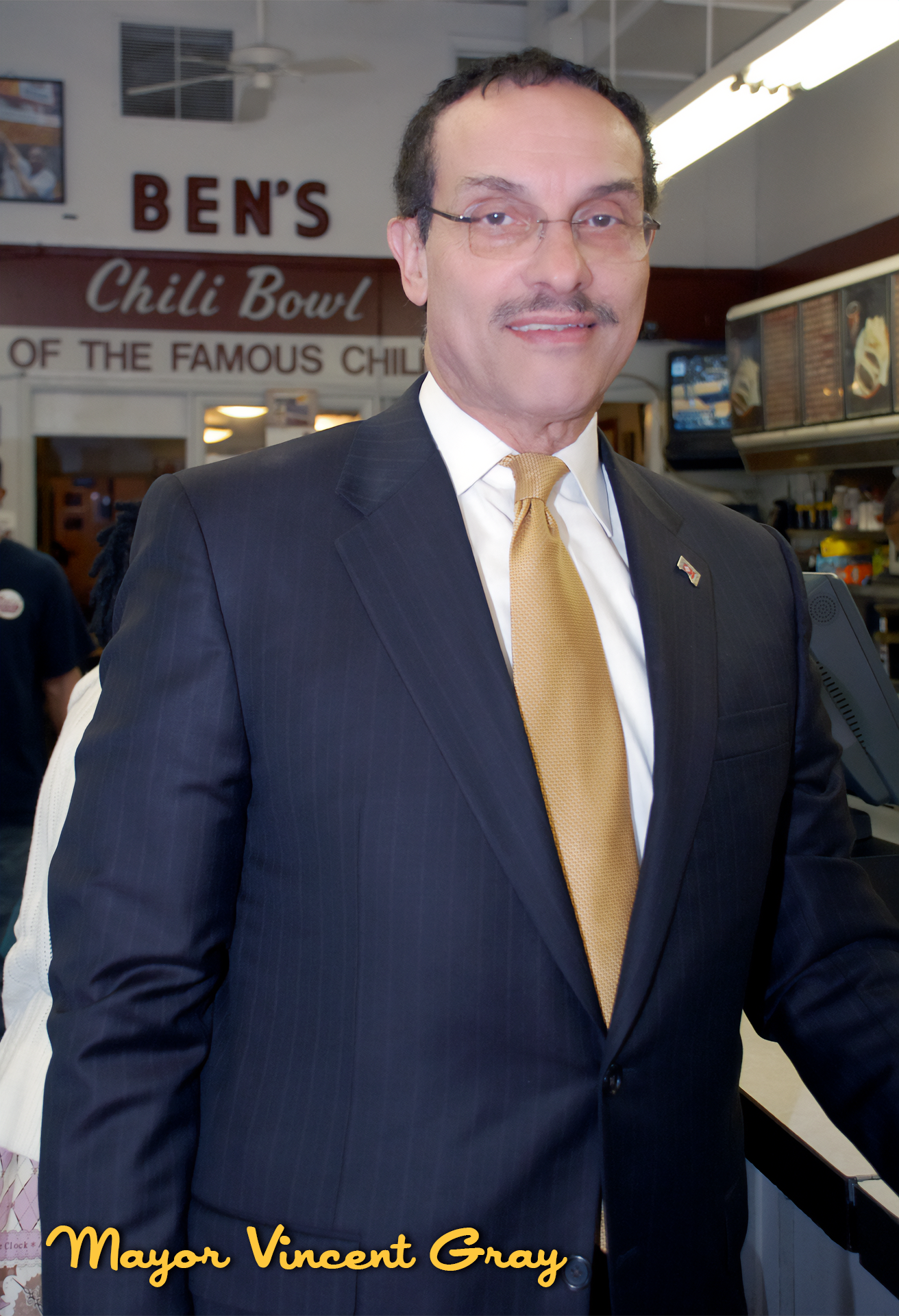 Mayor Vincent Gray standing inside a restaurant or diner, wearing a dark pinstripe suit and yellow tie, with a sign in the background reading "Ben's Chili Bowl" and "of the famous chili".