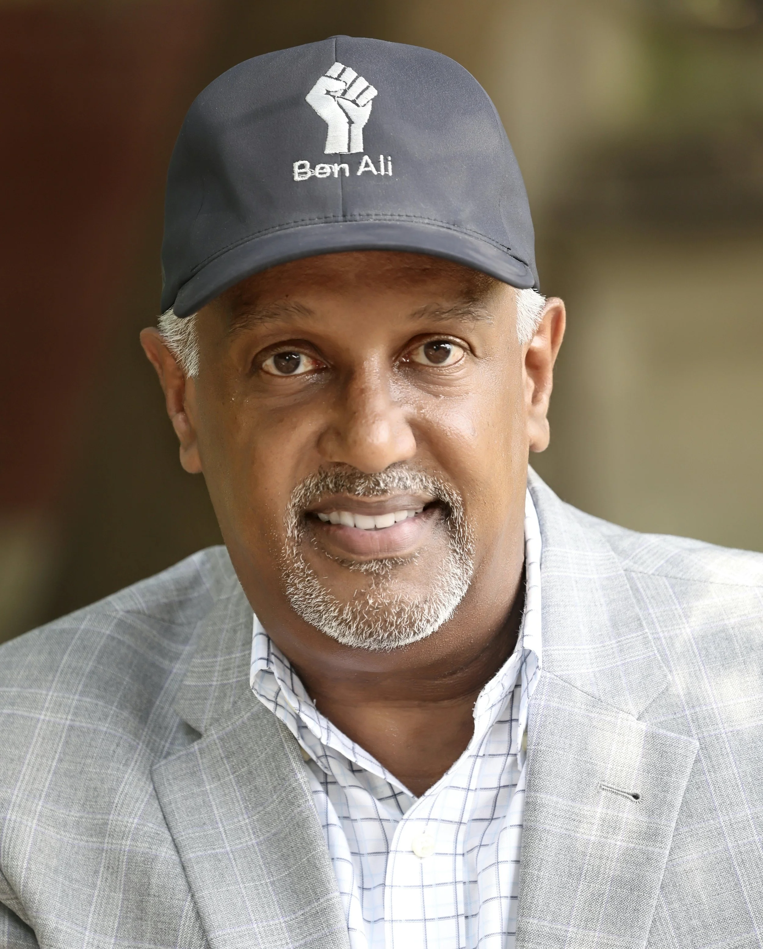 A man wearing a gray blazer and a white checkered shirt, with a gray goatee, wearing a navy blue cap embroidered with a white fist and the name 'Ben Ali'.