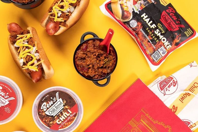 Hot dogs with shredded cheese, chili, and onions, a bowl of chili, a hot dog bun box, and hot dog chili sauce containers on a yellow surface.