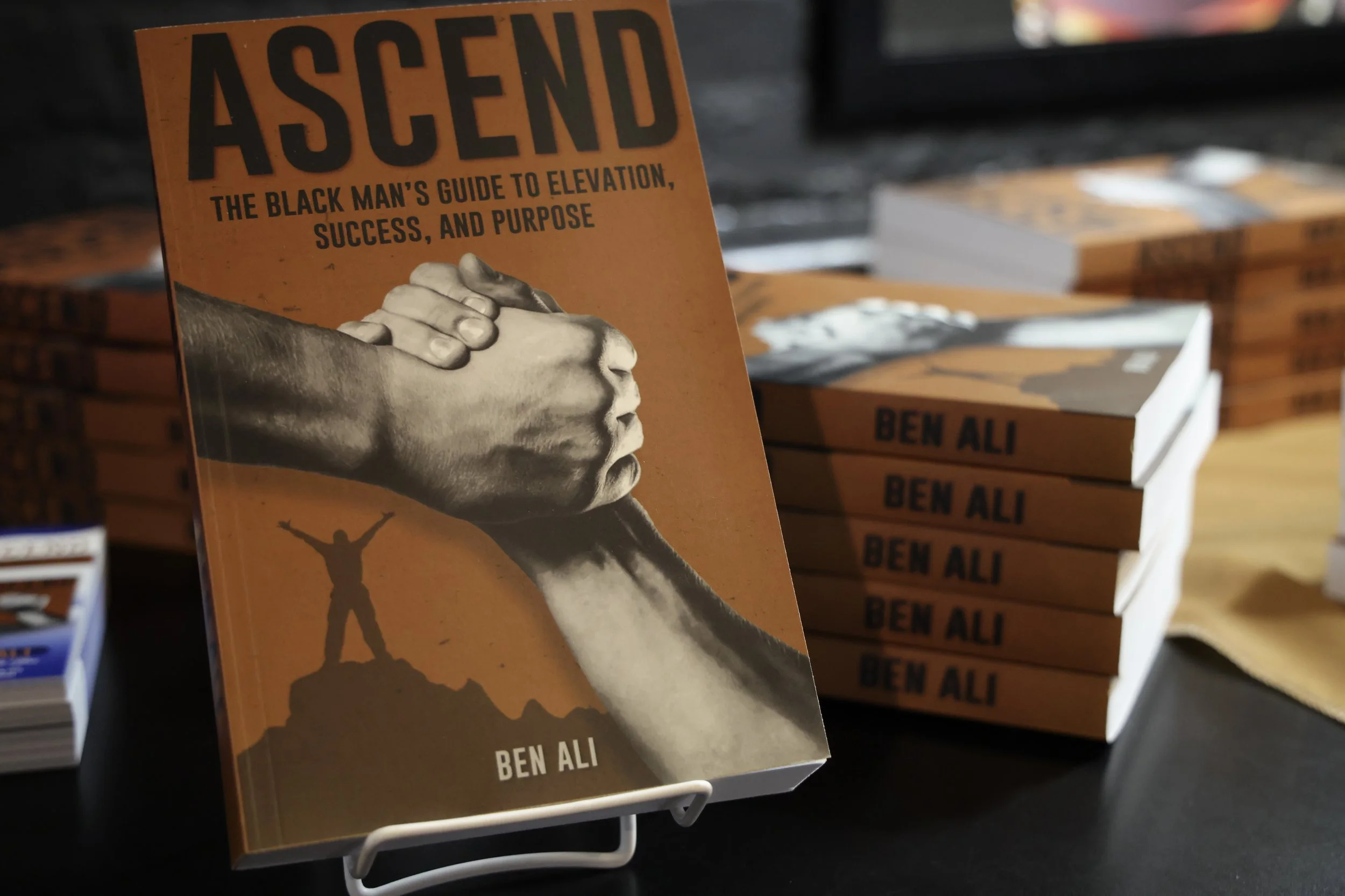 Book titled 'Ascend: The Black Man's Guide to Elevation, Success, and Purpose' with a cover illustration of two hands gripping each other, one hand pulling the other up, and a small figure standing on a peak with arms raised.