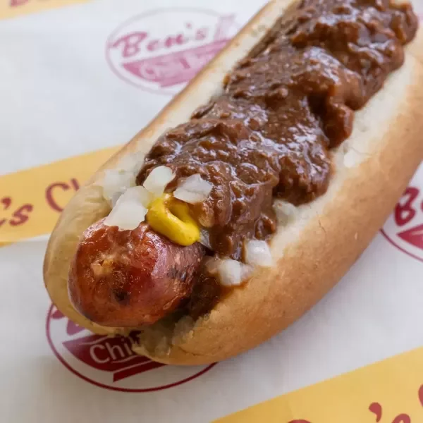 Hot dog with sausage, topped with chili, chopped onions, and mustard, in a bun.