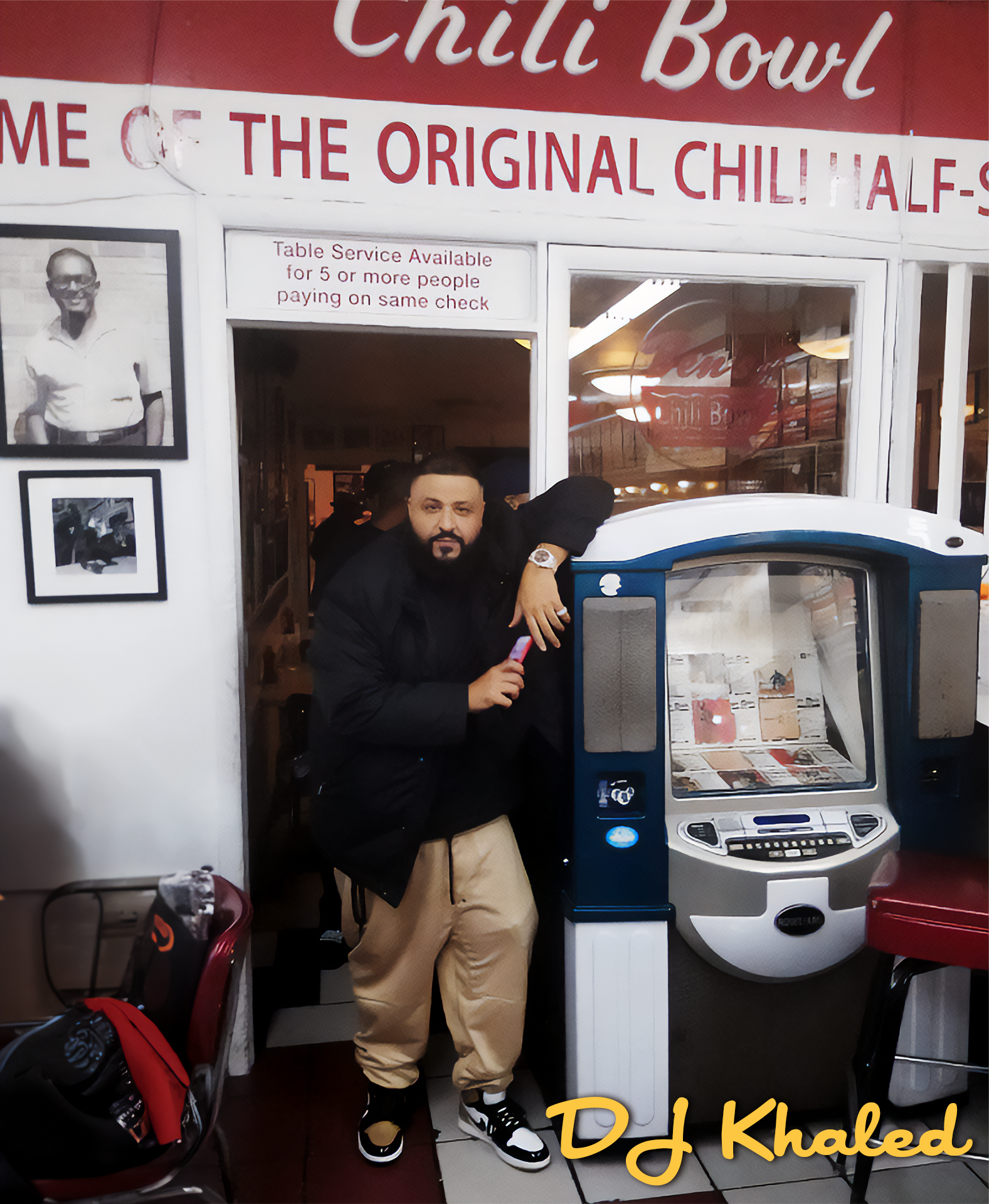 A man with a beard and black hair, wearing a black jacket, beige pants, and sneakers, leans on a vending machine in a restaurant called Chilli Bowl. The restaurant has a red sign with white text above the entrance, and pictures on the wall. The man i