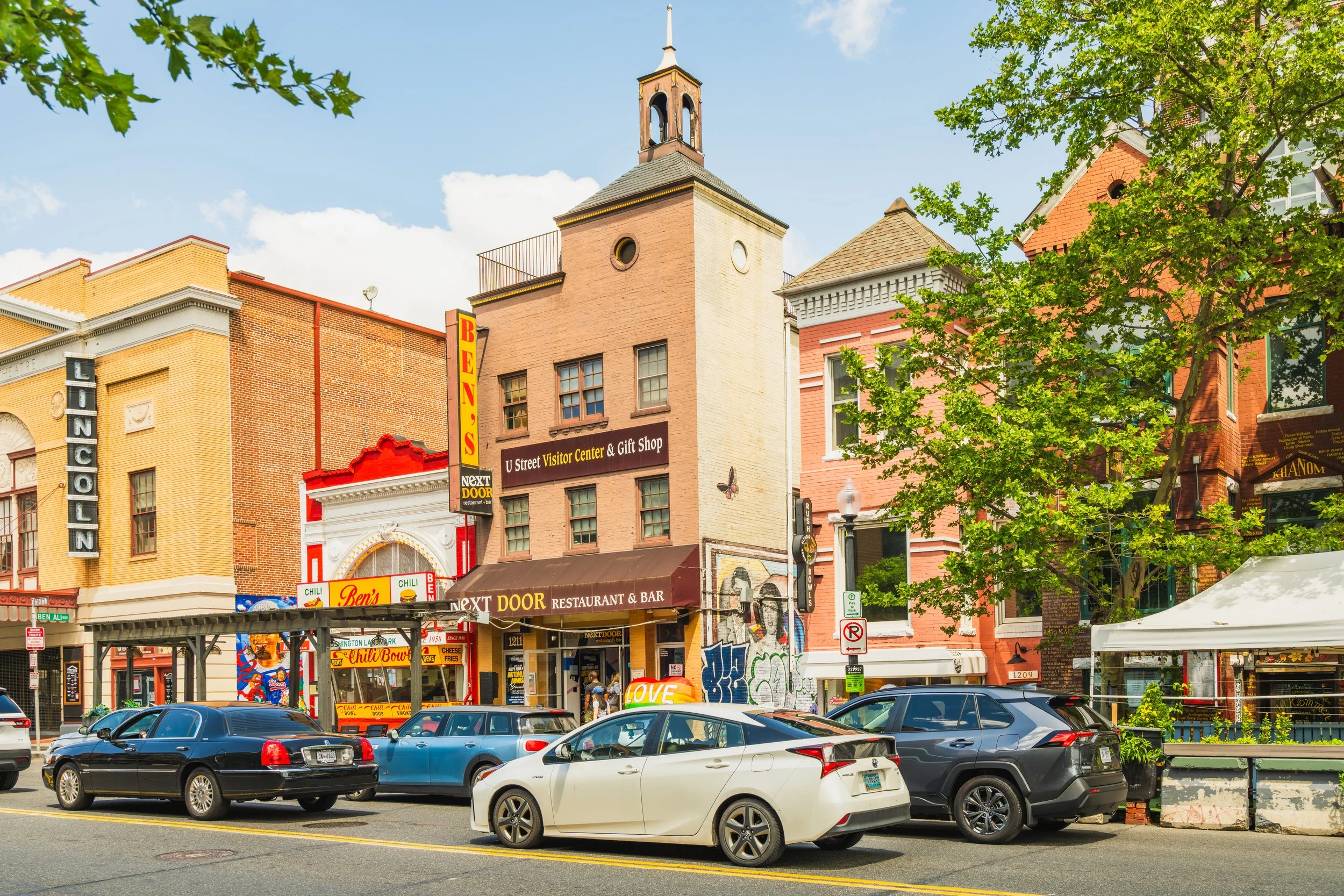 Street view featuring colorful buildings, parked cars, and a tree with green leaves in front of a restaurant and shops.