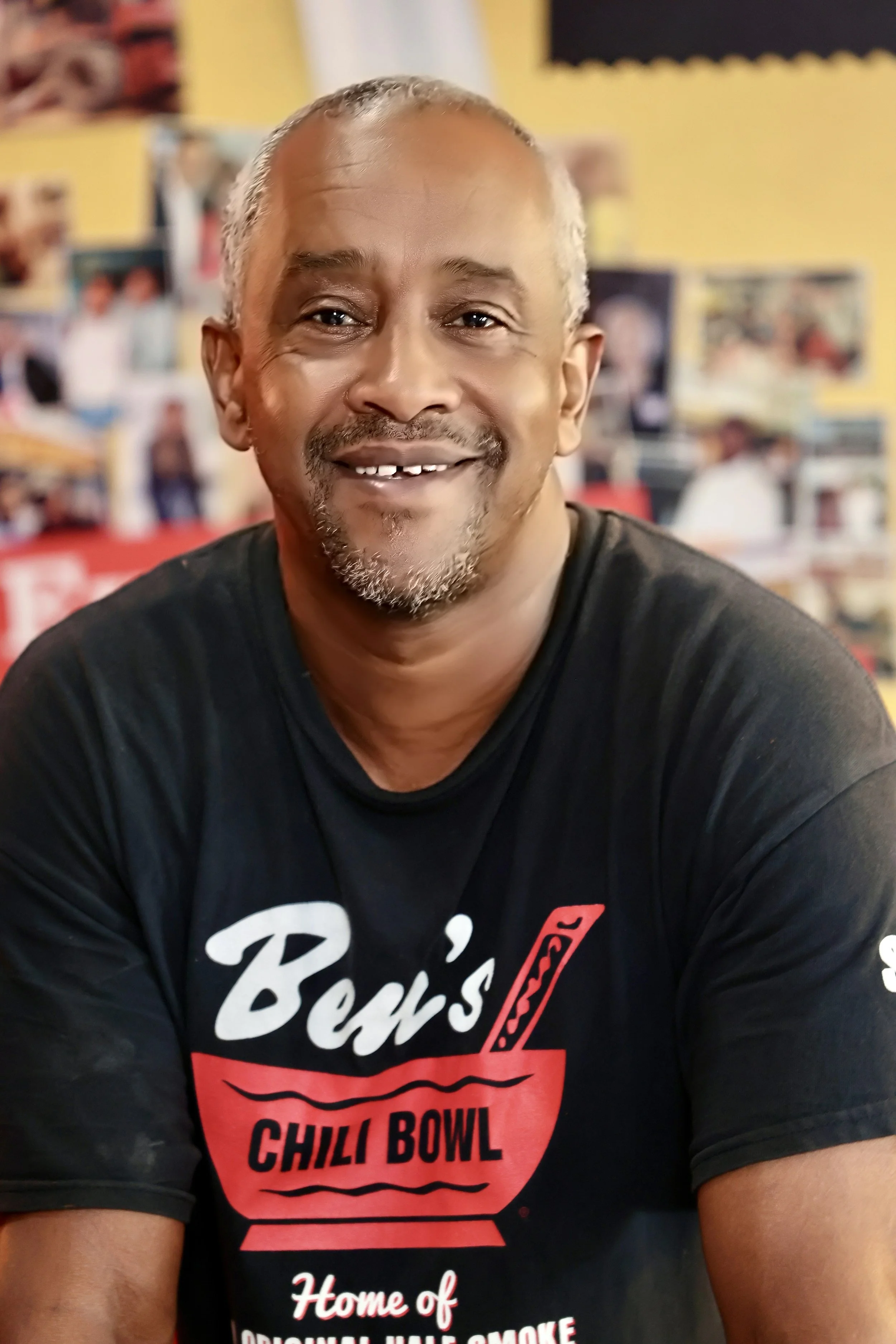 A smiling man with a goatee and short gray hair, wearing a black T-shirt with a logo and text, sitting indoors with a blurry background of photos on a wall.