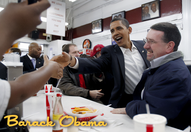 Barack Obama shaking hands with a man at a diner counter, surrounded by other people, smiling, with framed photos hanging on the wall behind them.