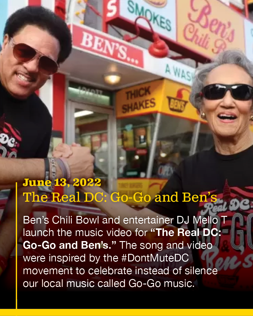 Two people, a man with dark hair and sunglasses and a woman with gray hair and sunglasses, smiling and shaking hands in front of a food stand with signs for Ben's Chili Bowl, thick shakes, and smoked foods.