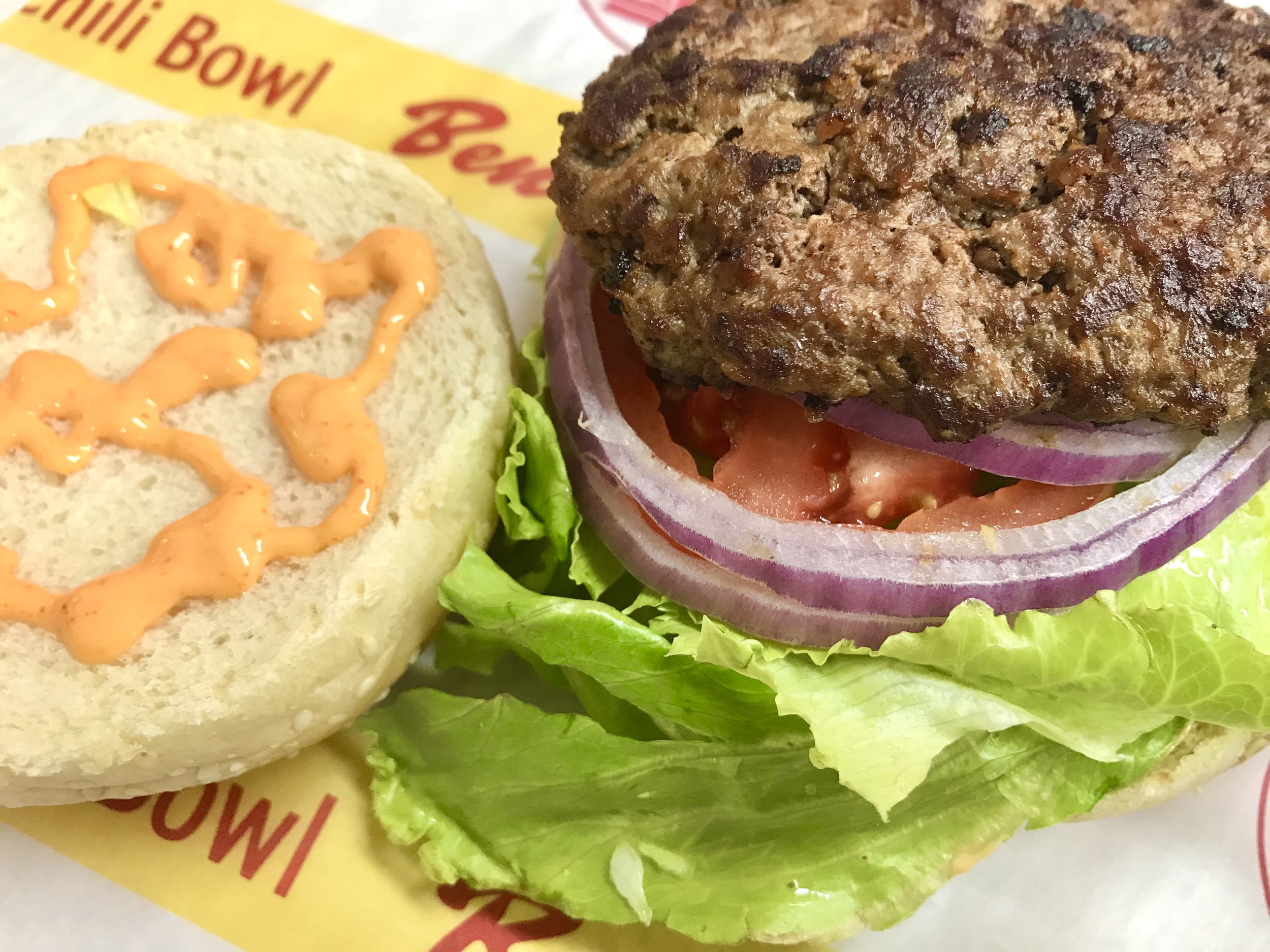 Close-up of a hamburger with lettuce, tomato, onion, and a beef patty on a bun. The sandwich is on paper with red and yellow printing.