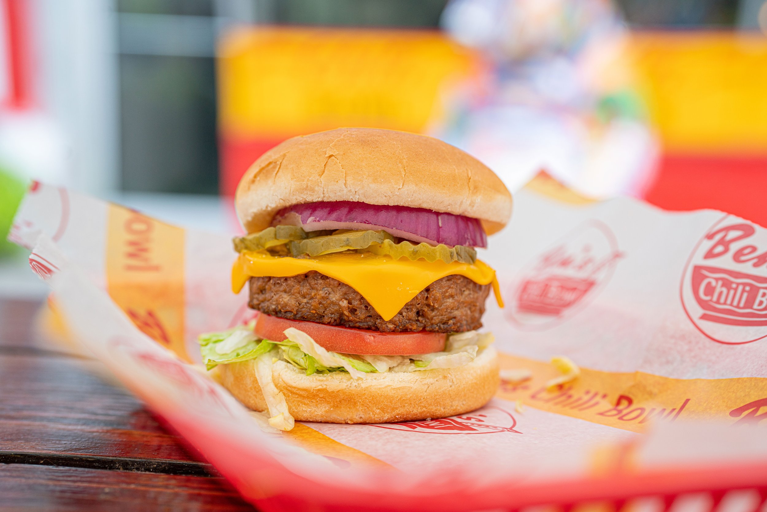 A cheeseburger with pickles, onions, tomato lettuce, cheddar cheese, hamburger patty, served in a bun on branded paper wrapping.