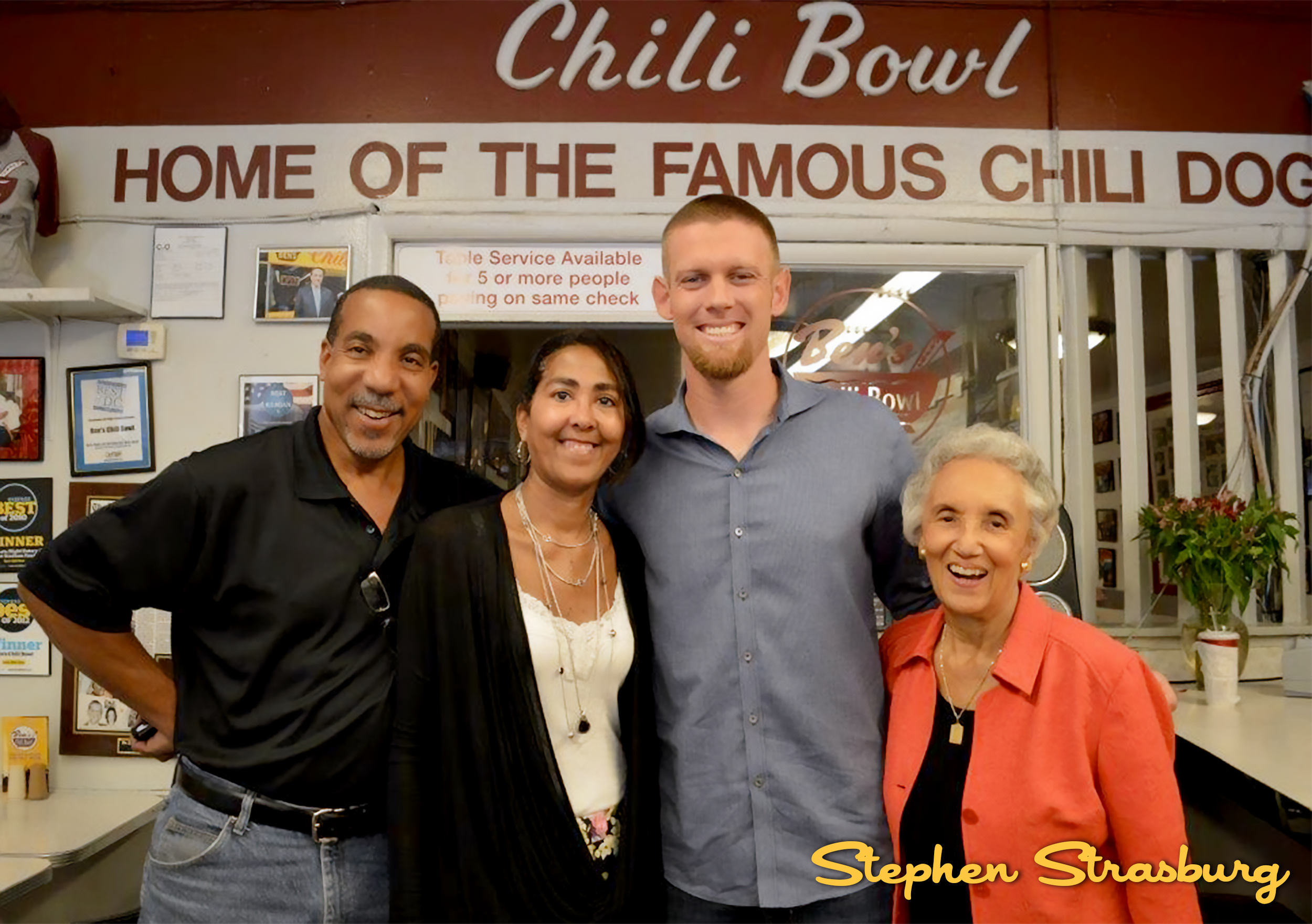 Four people smiling in front of a sign that says 'Chili Bowl' and 'Home of the Famous Chili Dog' inside a restaurant. The group includes two men and two women, one of whom is elderly. There are checks, awards, and a potted plant visible in the backgr