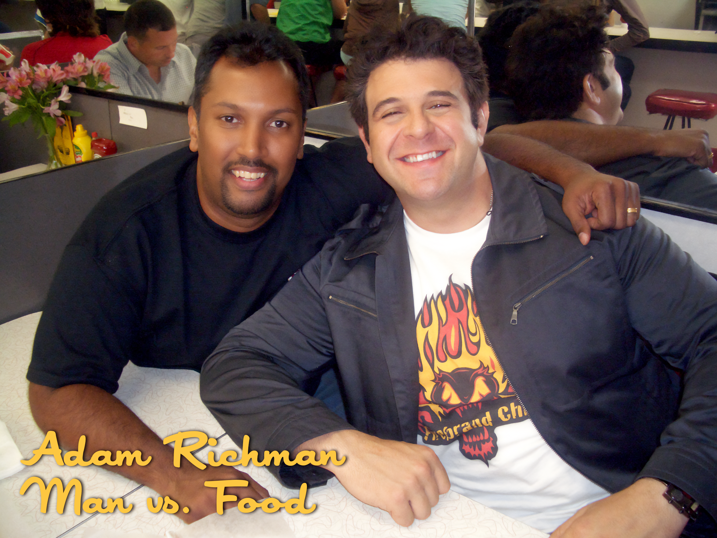 Two men smiling and sitting at a restaurant table, with one man having his arm around the other. The man on the left is wearing a black shirt, and the man on the right is wearing a black leather jacket and a graphic T-shirt with a fire and lion desig