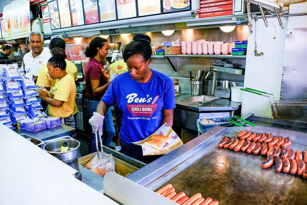 Workers at Ben's Chili Bowl restaurant cooking hot dogs on a grill, with customers in the background ordering food.