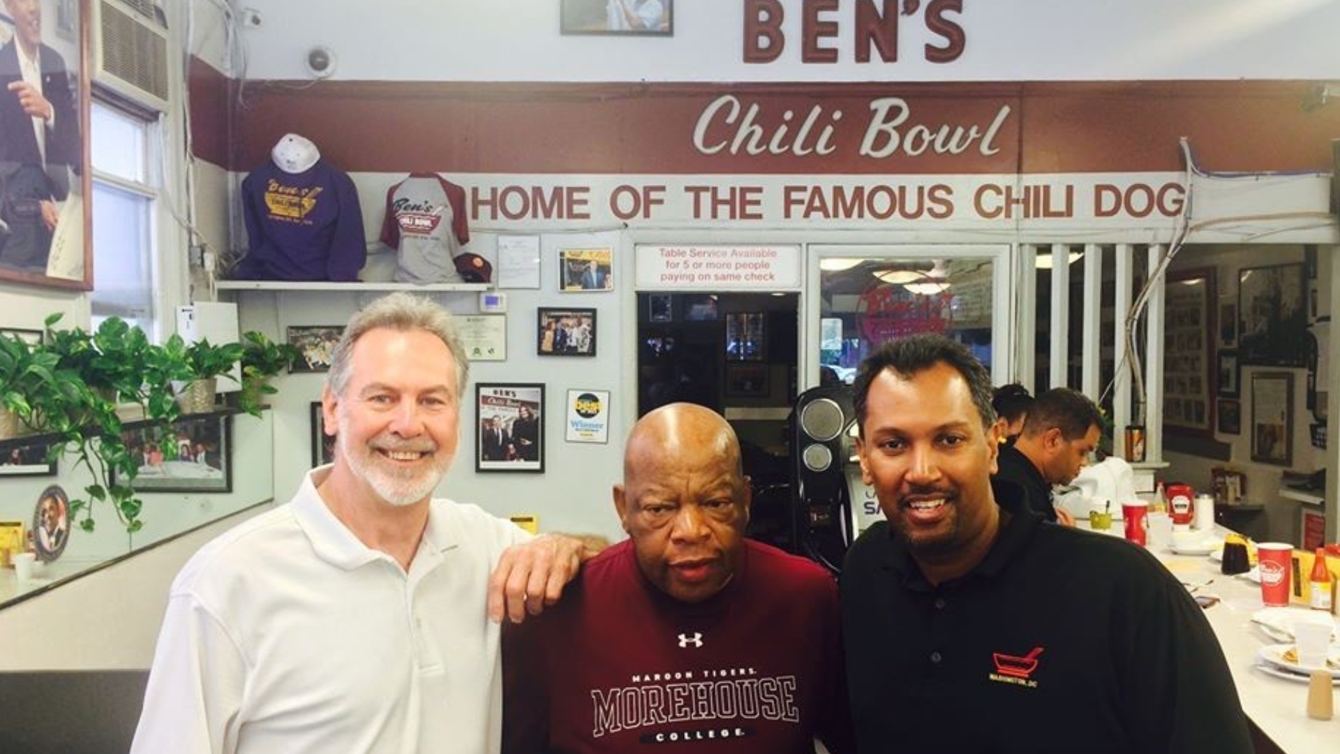 Three men stand inside a restaurant with a sign above them that says "Ben's Chili Bowl." The man on the left has gray hair and a beard, wearing a white shirt. The man in the middle is bald and wears a maroon shirt with college name "Morehouse." The man on the right has dark hair and wears a black polo shirt. The restaurant has framed pictures on the wall and seating in the background.