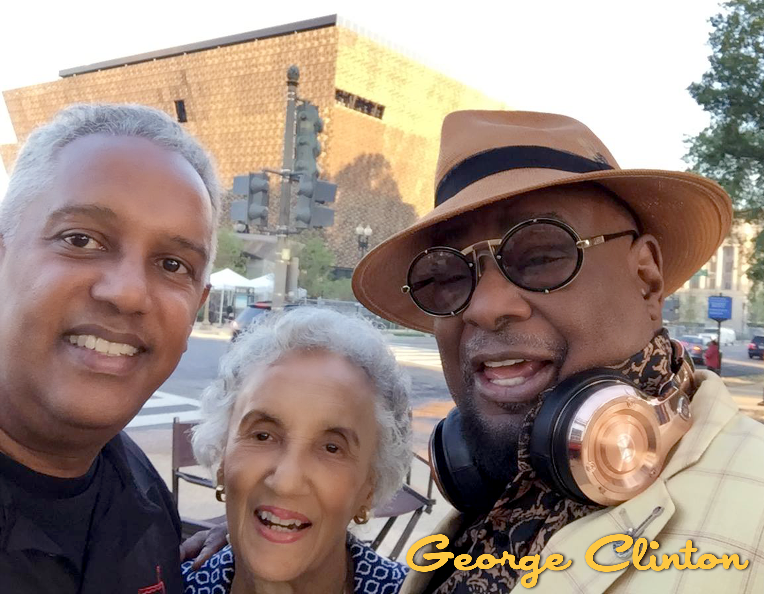 Three people, two men and one woman, smiling for a selfie outdoors with a city street and brick building in the background. The man on the right is wearing a tan hat, round sunglasses, and has headphones around his neck. The woman has gray hair and i