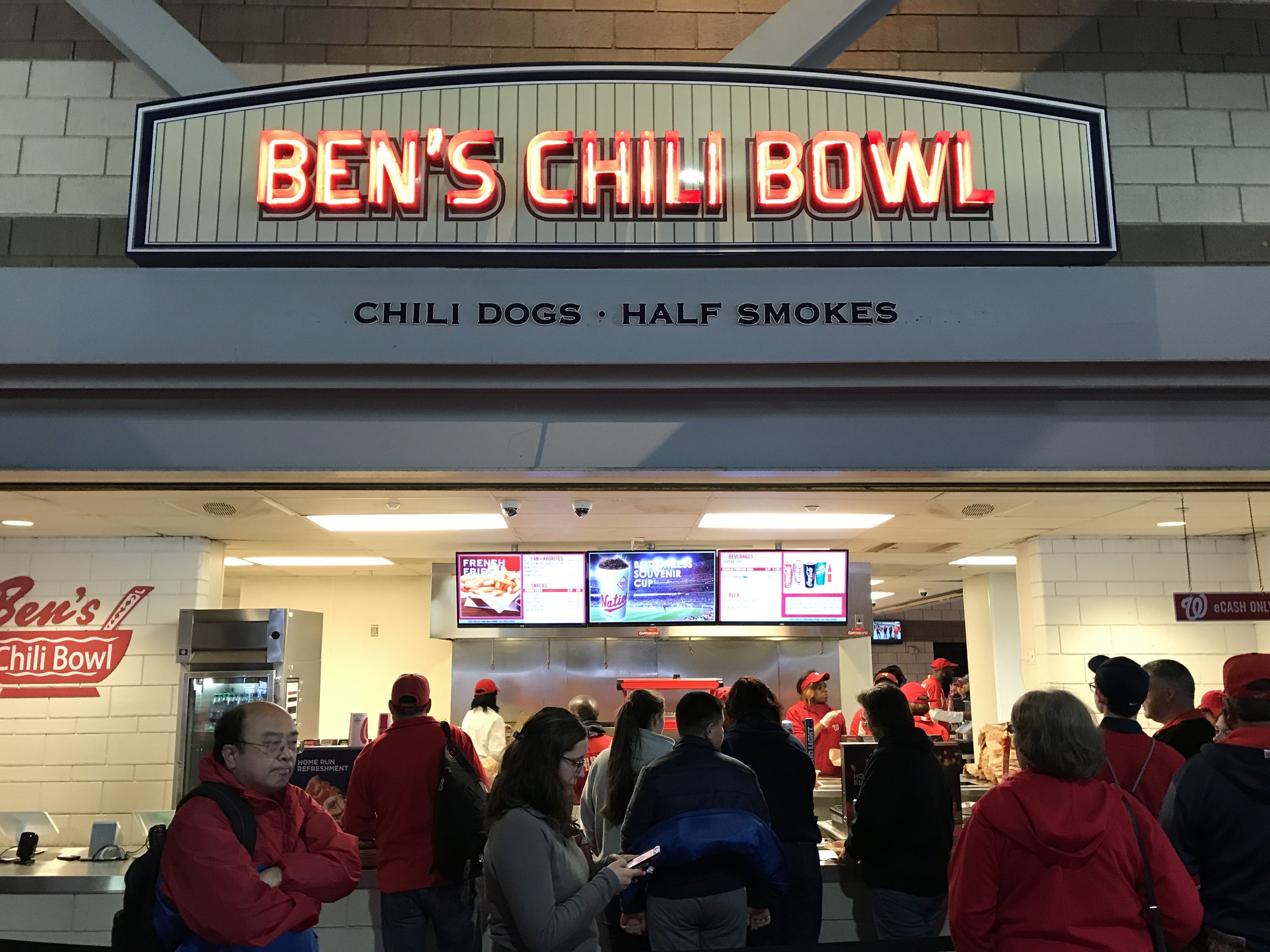 People in line and at the counter of Ben's Chili Bowl restaurant, with a lit sign above reading 'Ben's Chili Bowl' and a menu board inside.