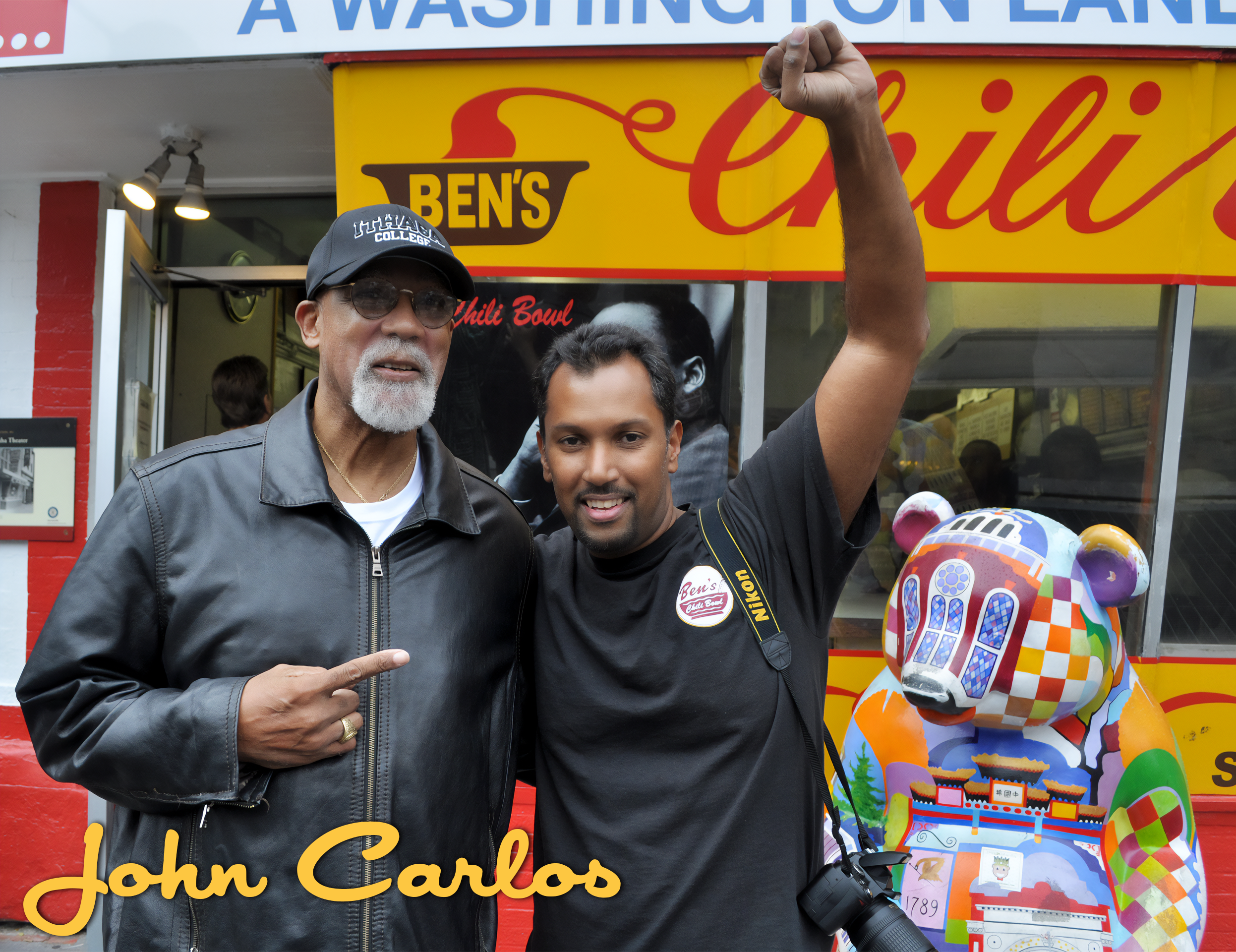 Two men standing in front of a bright yellow and red Ben's Chili Bowl food stand; one man has gray hair, a beard, glasses, and is wearing a black leather jacket and cap, while the younger man has short black hair, and is raising his right arm with a 