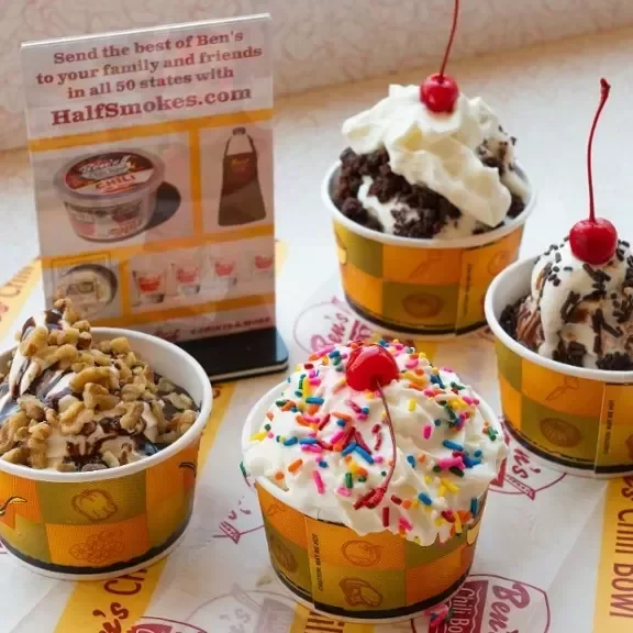Four cups of ice cream with various toppings on a table, with a promotional sign in the background.