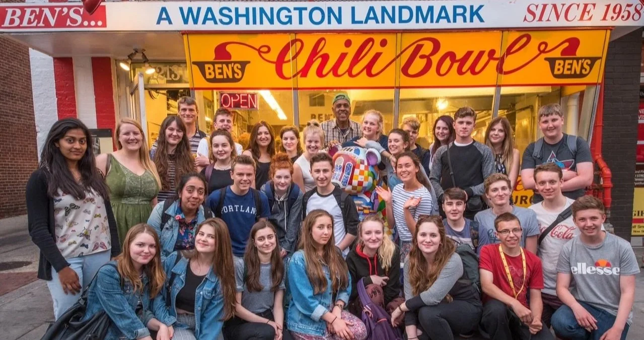 A large group of teenagers and some adults standing outside a restaurant called Ben's Chili Bowl, posing for a photo. The restaurant has a yellow and red sign, with an open sign illuminated in the window. The group appears happy and excited.