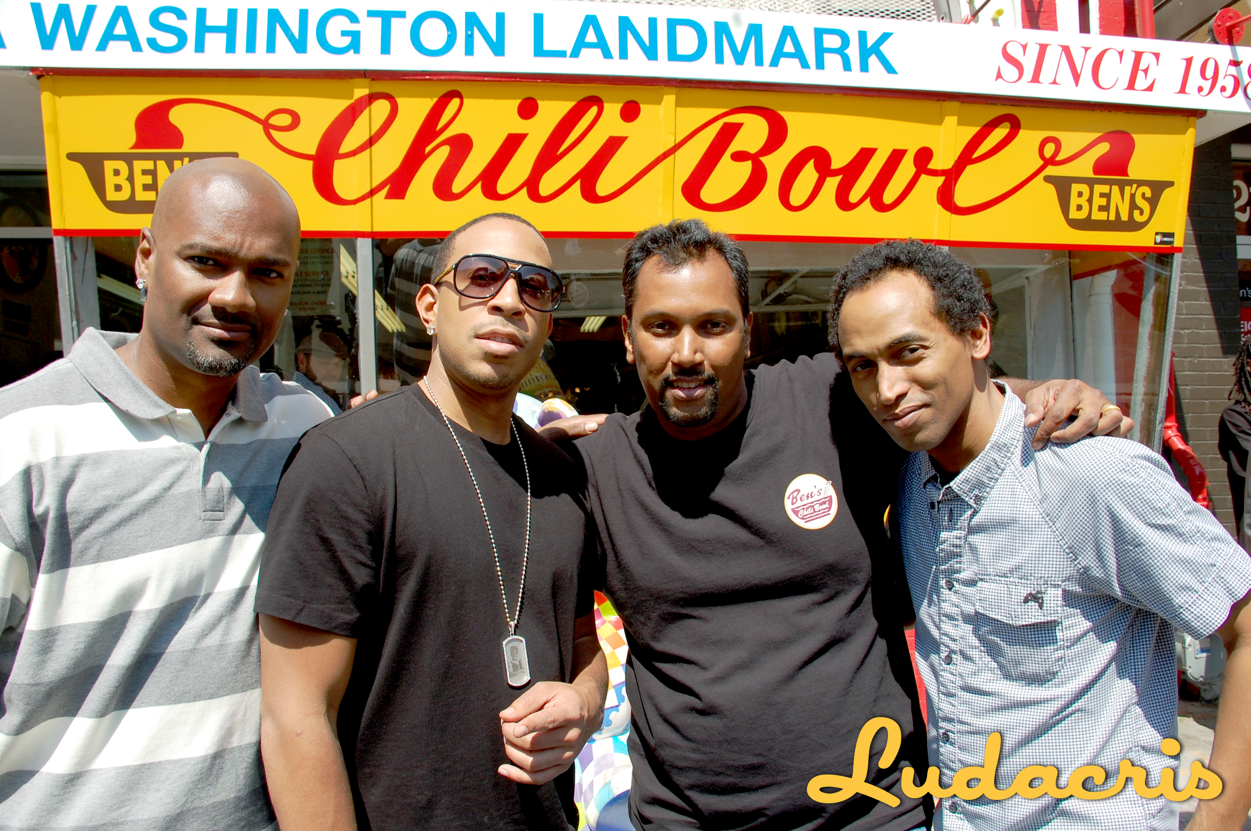 Four men standing together in front of a food stand labeled 'Chili Bowl' with a sign above that reads 'Washington Landmark since 1958'.