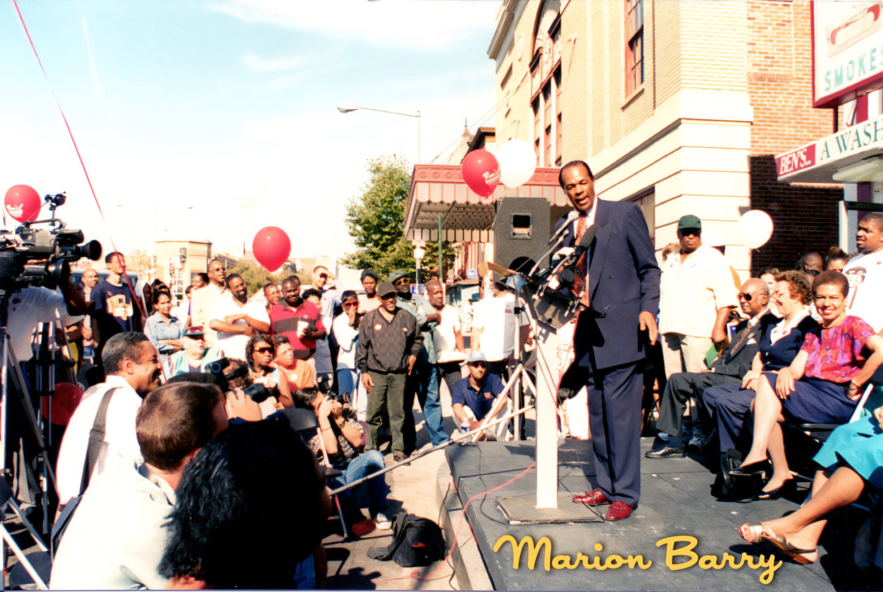 Marion Barry speaking at a podium during a public event outdoors, with a crowd of people, some seated and others standing, watching him. Red and white balloons are visible, and there is a camera crew recording the event. The setting features brick bu