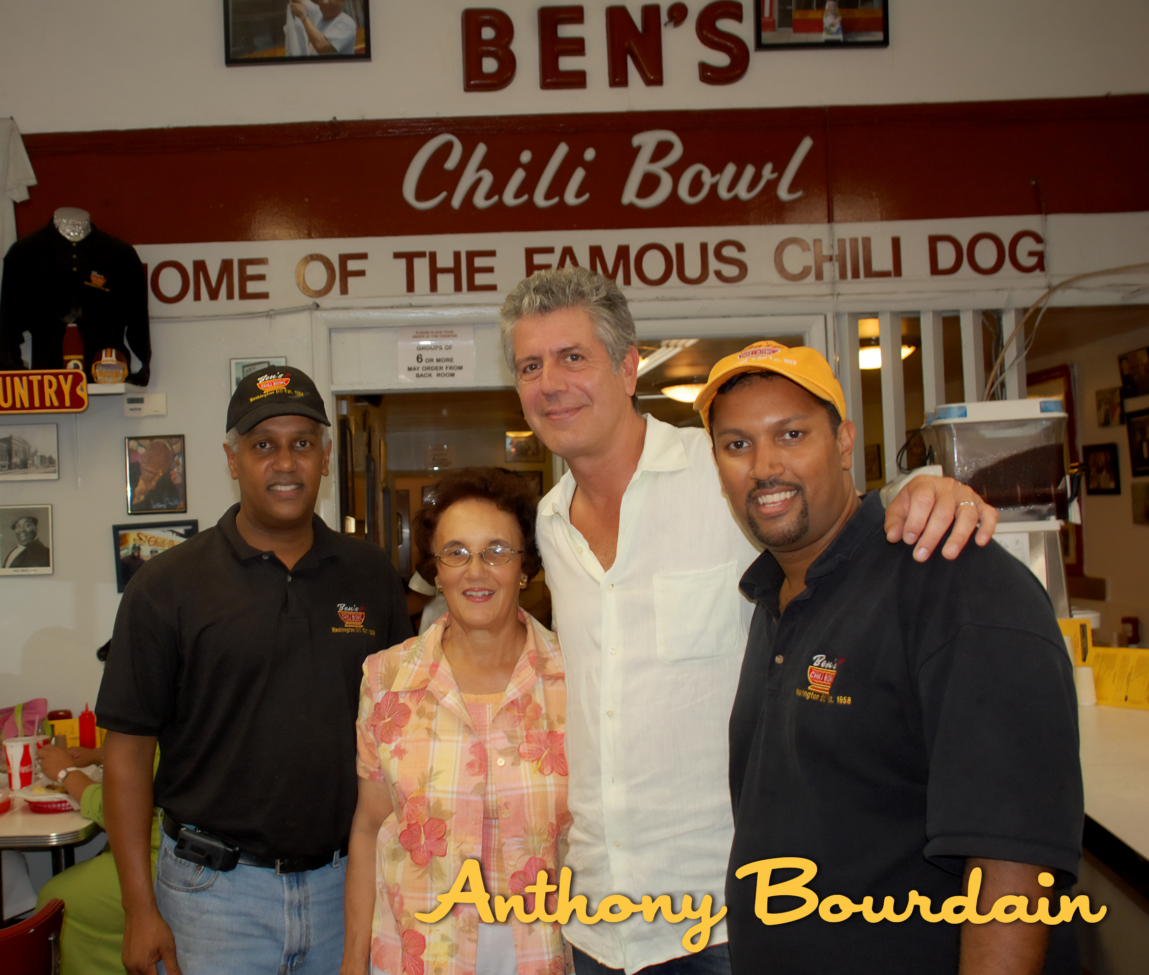 Four people standing inside Ben's Chili Bowl restaurant, smiling for the photo. The background features a sign that reads 'Home of the Famous Chili Dog' and various decorations on the wall.