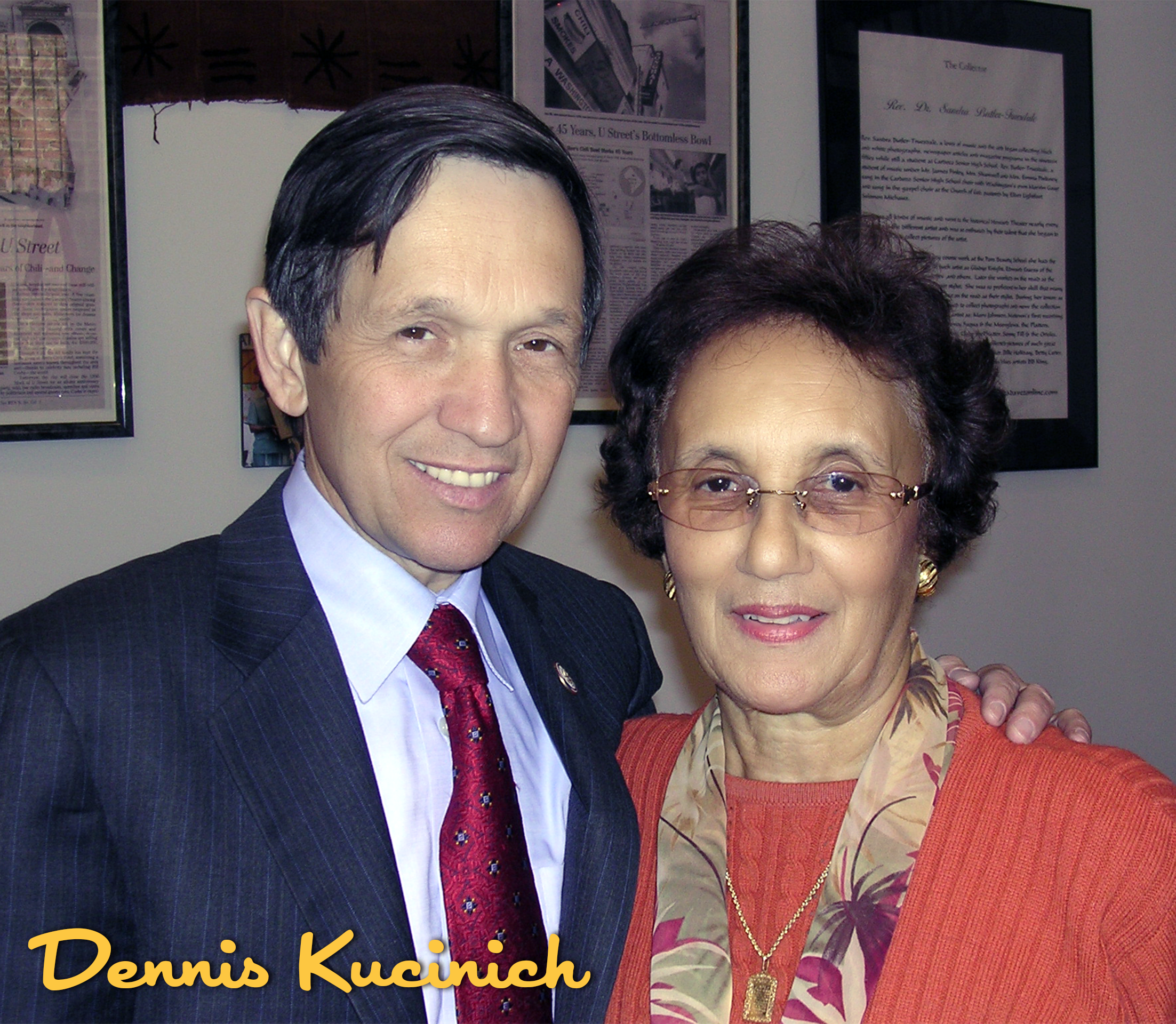 A man in a dark pinstripe suit and red tie, and a woman in an orange sweater with glasses, standing together indoors in front of framed documents on the wall. The man has his arm around the woman's shoulder. The image has the text 'Dennis Kucinich' i