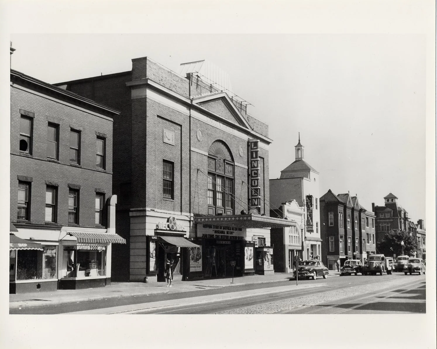 Black and white photograph of a vintage city street with theater and storefronts, cars parked along the curb, and pedestrians walking on the sidewalk.