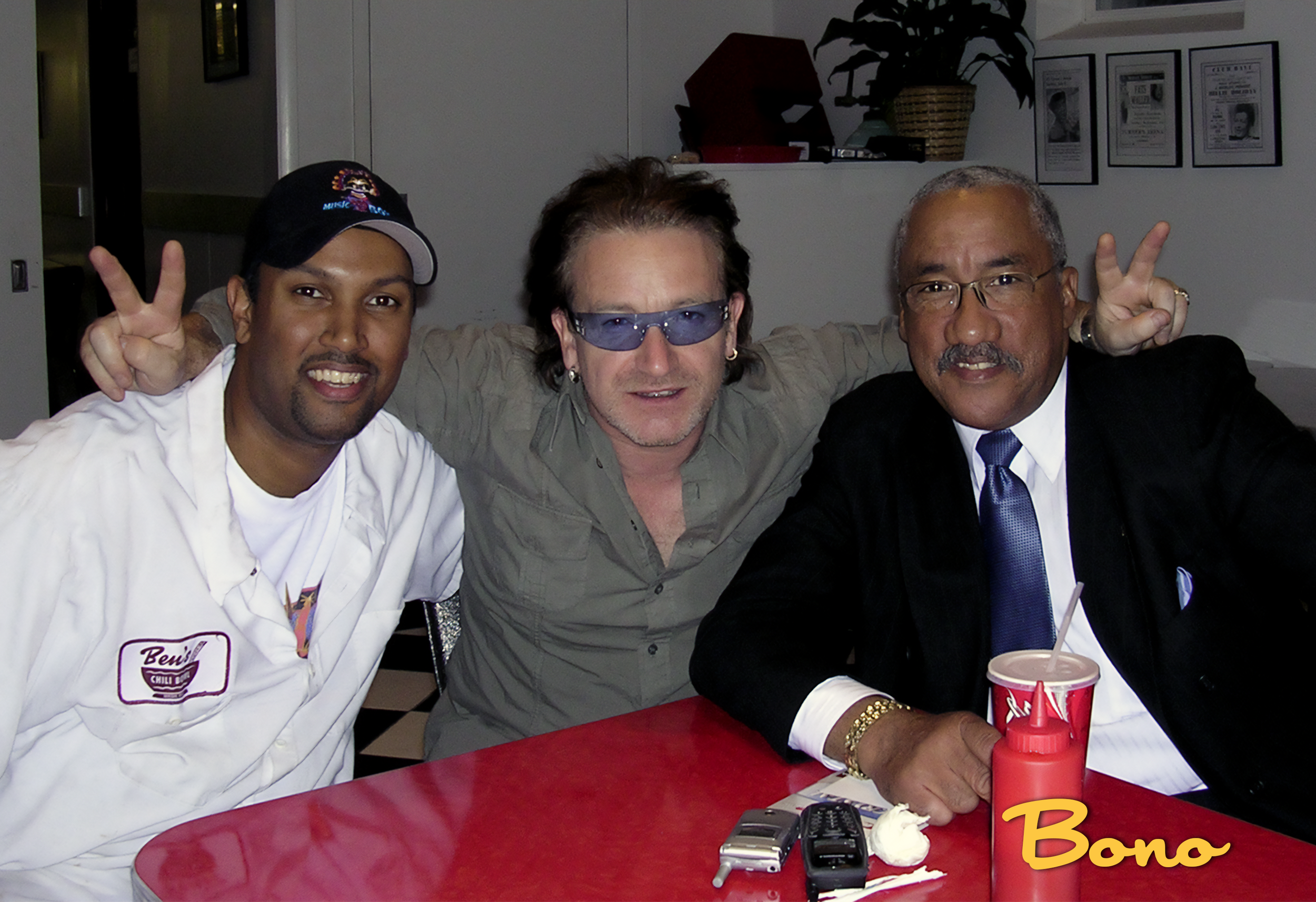 Three men sitting at a red table. The man on the left is wearing a white chef's coat and a black cap. The man in the middle has sunglasses and is dressed casually. The man on the right is wearing a black suit, white shirt, and a blue tie. They are al