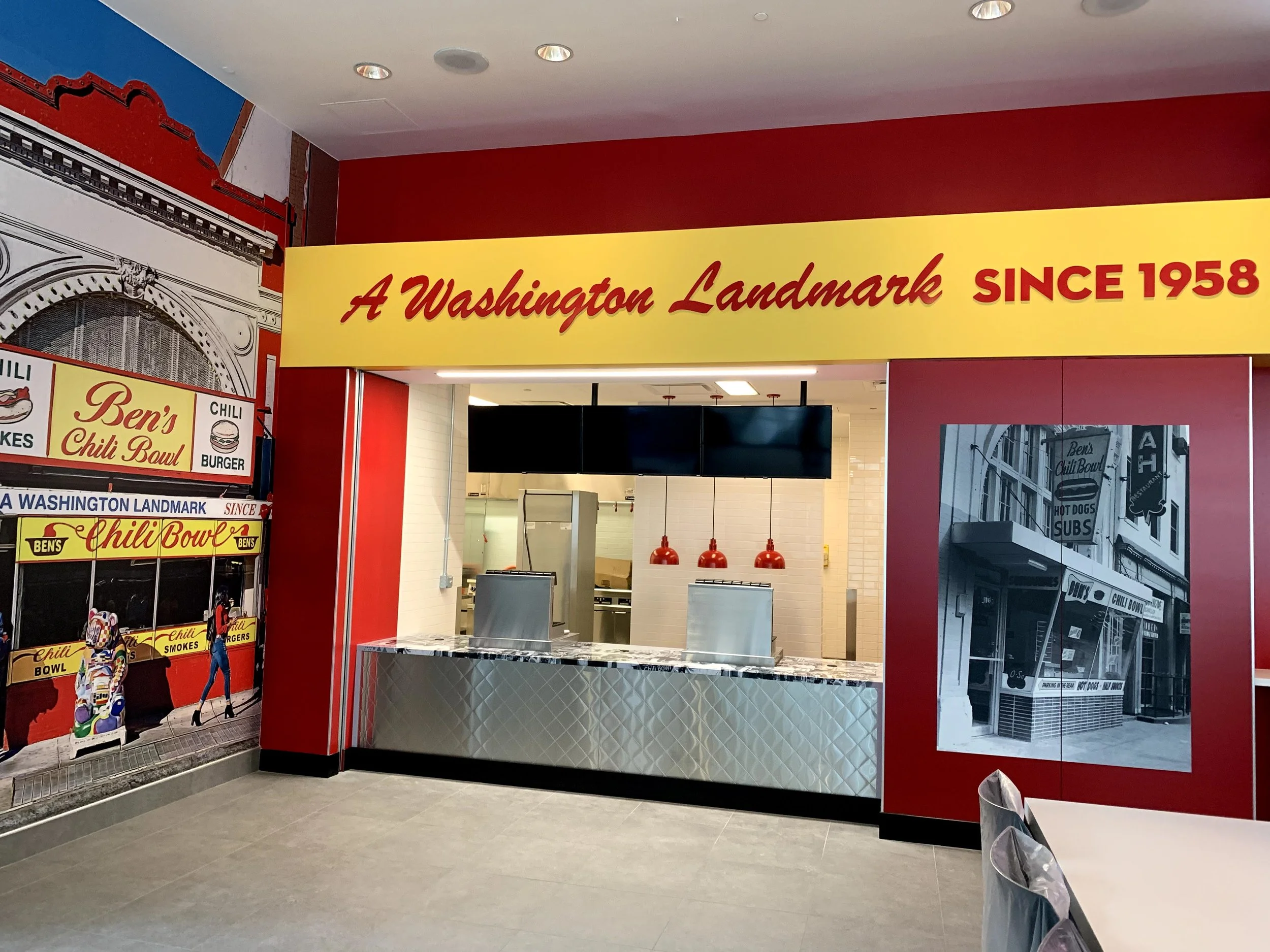 The interior of a restaurant with bold red and yellow signage stating, 'A Washington Landmark SINCE 1958.' Inside, food service counters and kitchen area are visible, with a black and white photograph of the restaurant's exterior on the right wall and a colorful mural of the same location on the left wall.