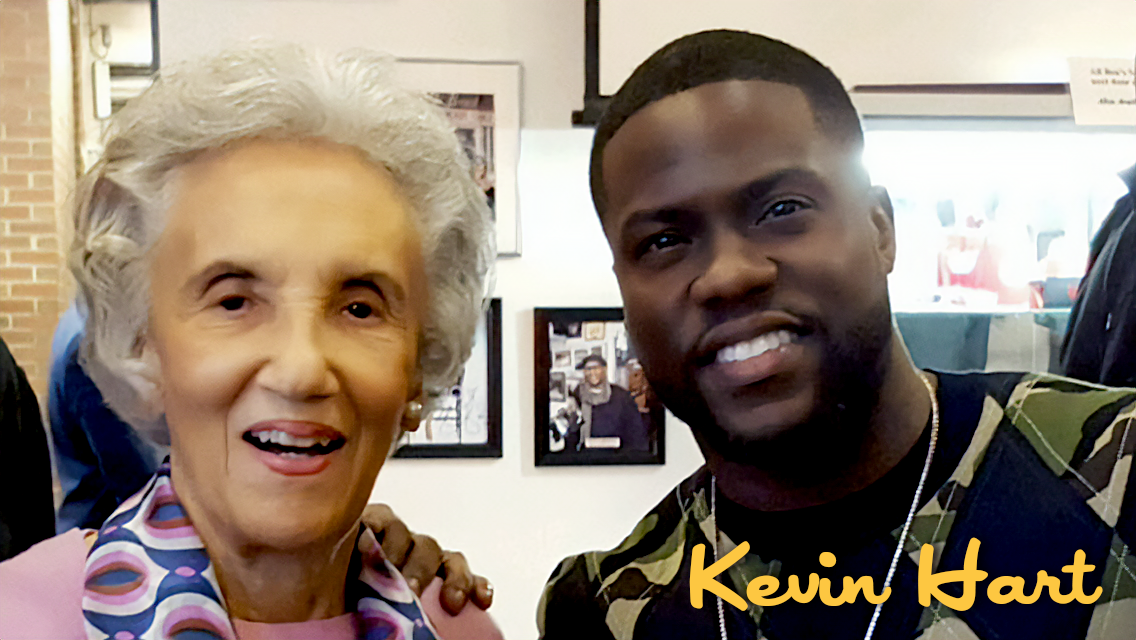An elderly woman with gray hair smiling next to a young man with short dark hair smiling. They are in an indoor setting, possibly a restaurant or cafe, with framed pictures on the wall in the background. The young man has a name tag that says "Kevin 