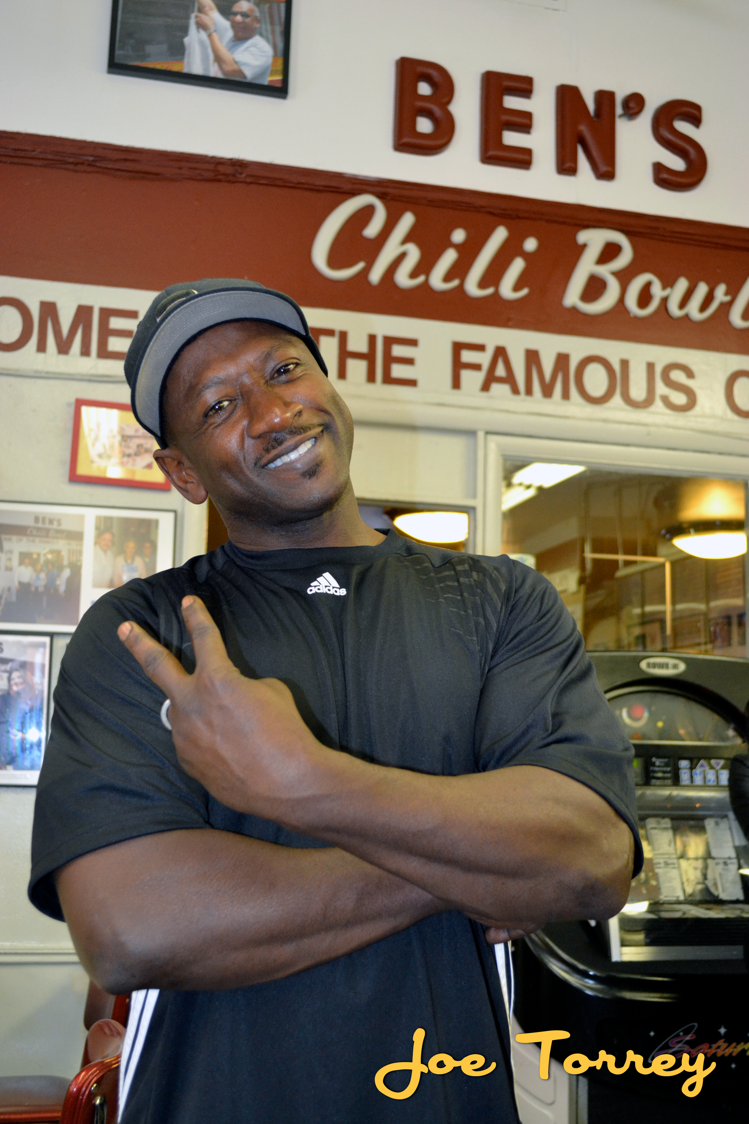 A man smiling and posing with a peace sign inside a restaurant with sign reading 'Ben's Chili Bowl.'