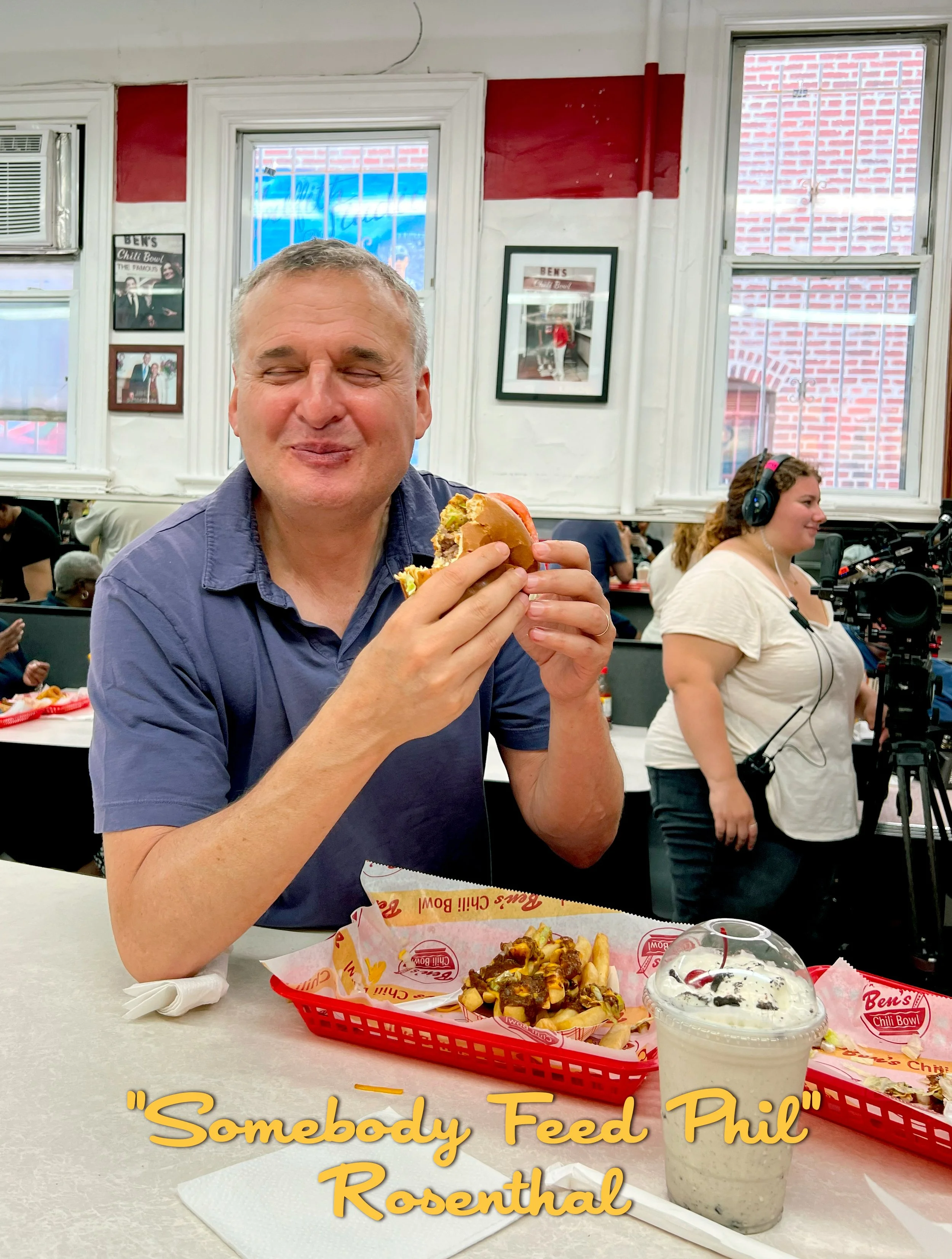 A man enjoying a burger at a casual restaurant, with a milkshake on the table, and a woman filming with a camera in the background.
