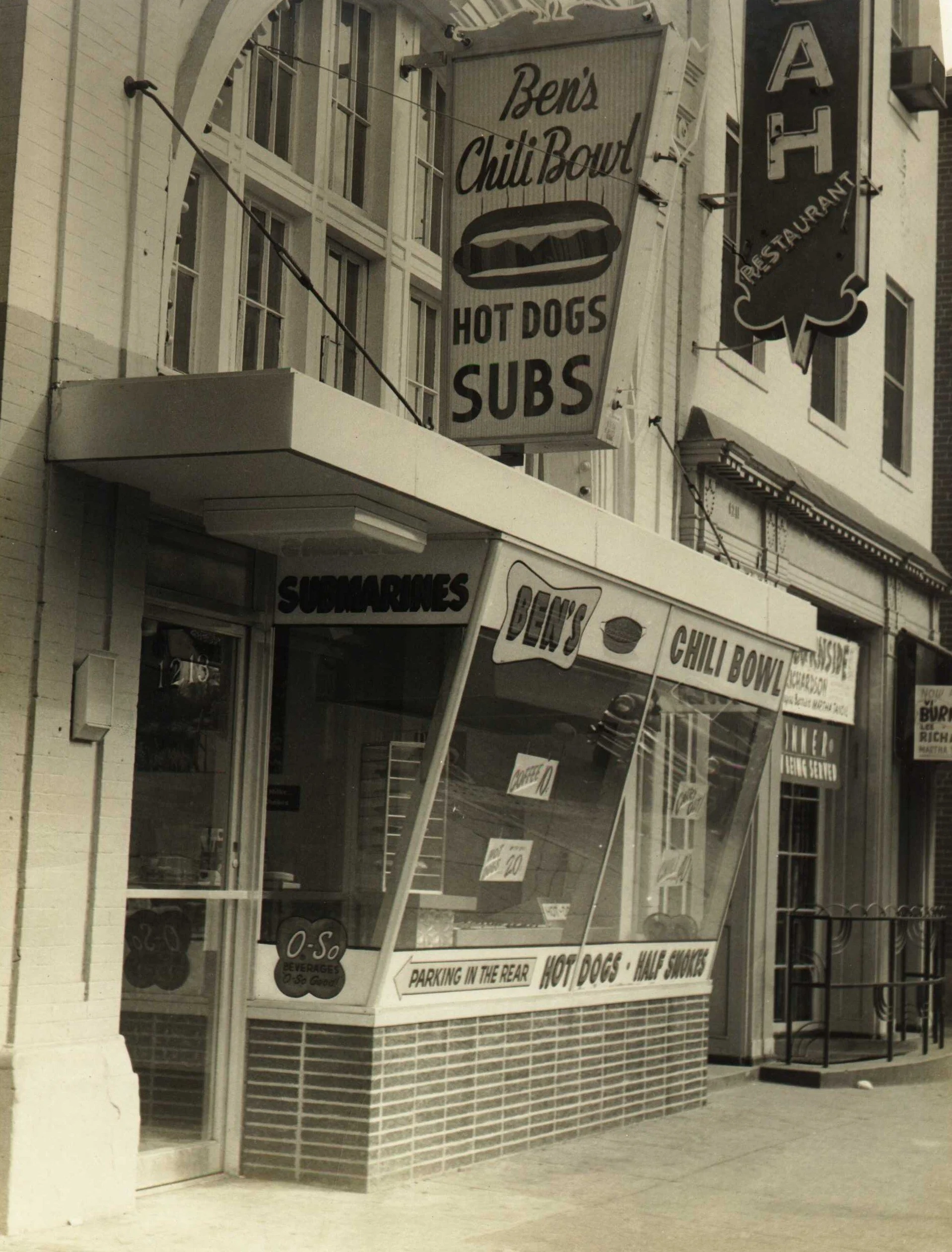 A small restaurant with signs advertising chili bowl, hot dogs, and subs, featuring a window service counter and storefront signage.