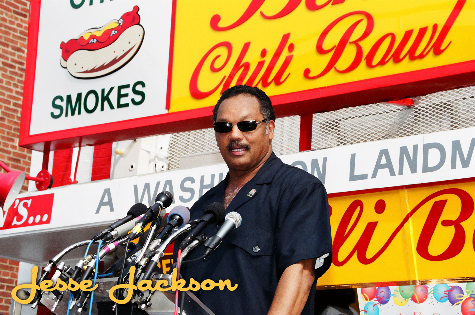 Jesse Jackson speaking at a podium with multiple microphones, standing in front of a hot dog stand sign. The sign reads 'Smokes' and 'Chili Bowl' in red and yellow colors.