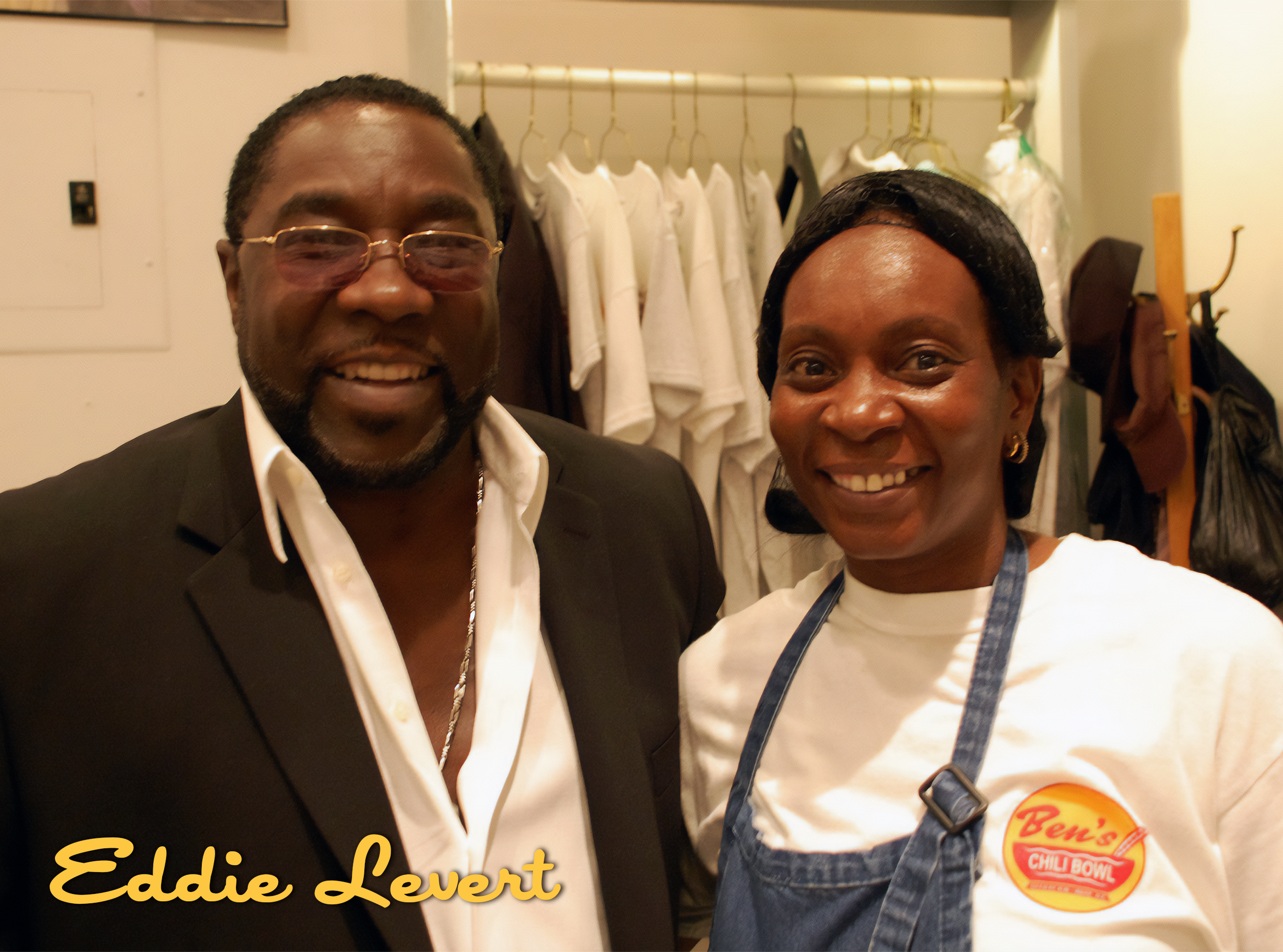 Man and woman smiling indoors, with clothing hanging in the background. The man is wearing glasses and a suit, while the woman is in a white shirt and apron with a Ben's Chili Bowl logo.