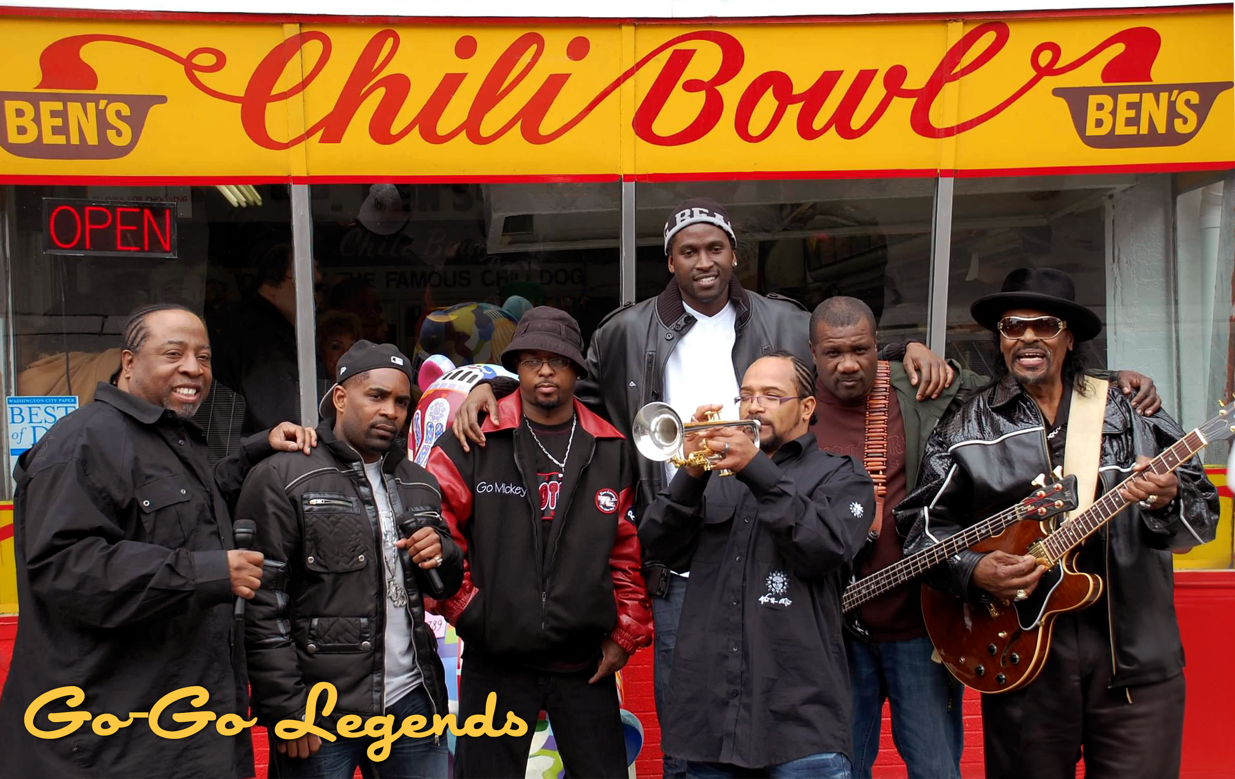 A group of seven men standing in front of a food truck named Ben's Chilli Bowl, some holding musical instruments and others with microphones, with the words 'Go-Go Legends' on the bottom left corner.