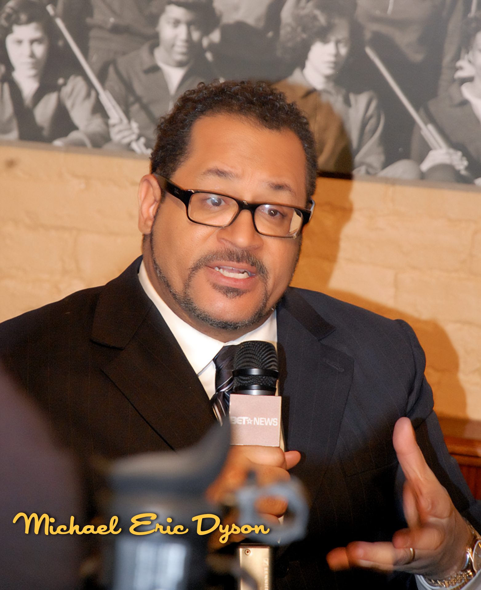 Michael Eric Dyson speaking into a microphone during an interview, wearing a black suit, white shirt, and striped tie, with a black-and-white photo of young men in the background.