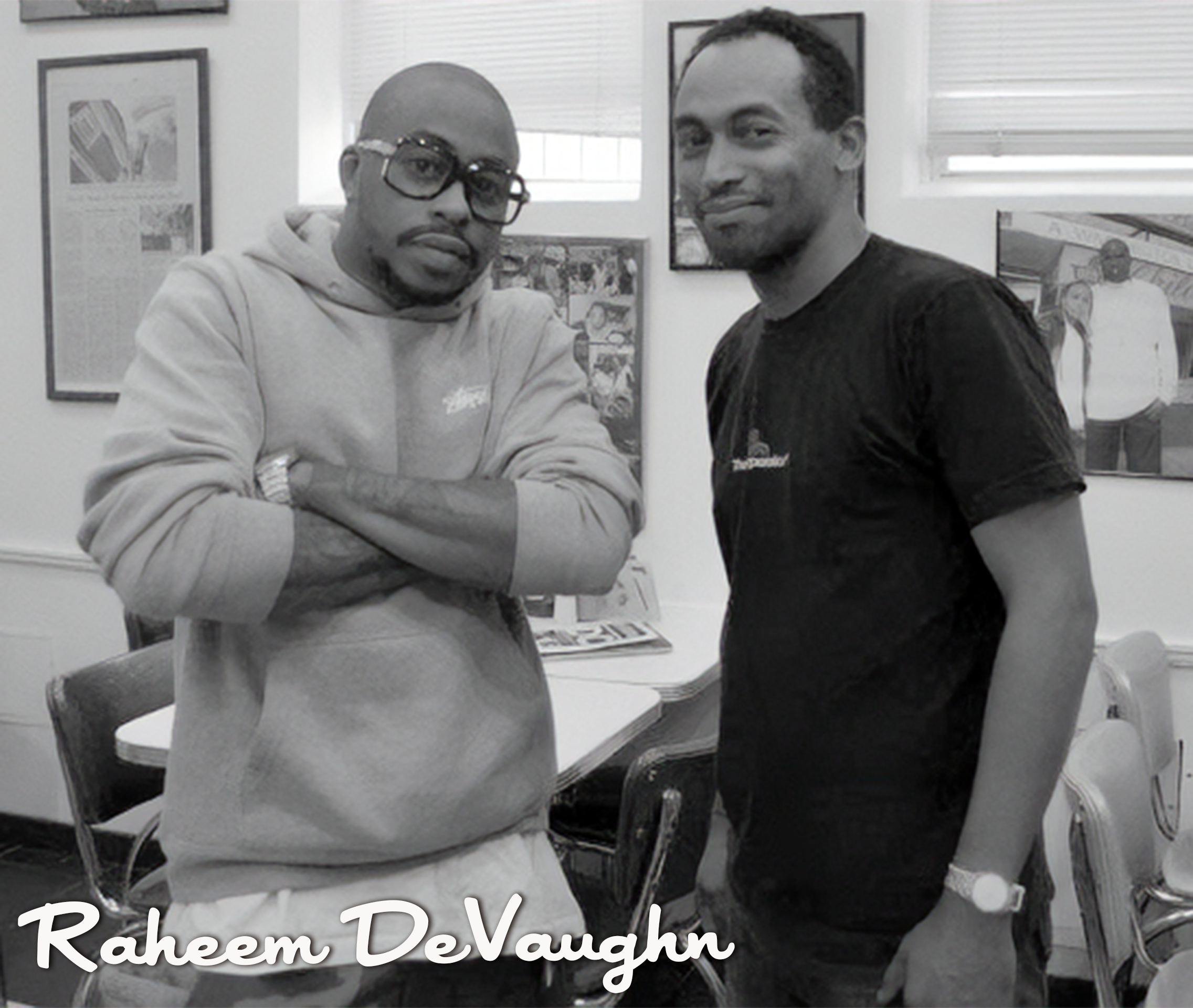Two men standing in an indoor setting, one with arms crossed wearing glasses and a hoodie, the other wearing a black shirt with a watch on his wrist. They are posing for the photo with framed pictures on the wall behind them. The text 'Raheem DeVaugh