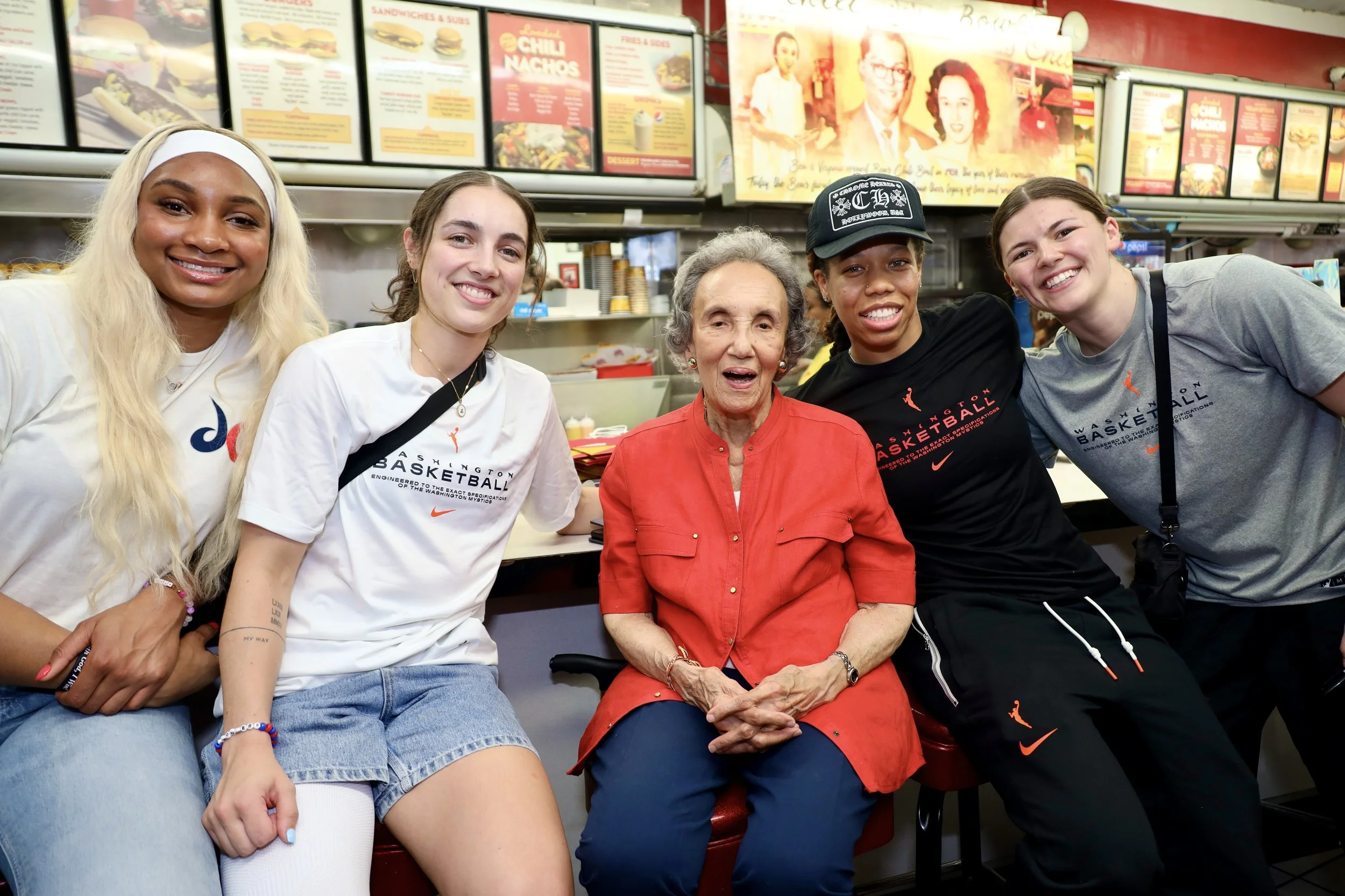 Group of four young women and one elderly woman sitting at a fast food restaurant counter, smiling for the camera. The young women are wearing casual sportswear, and the elderly woman is dressed in a red shirt. The background features menu boards wit