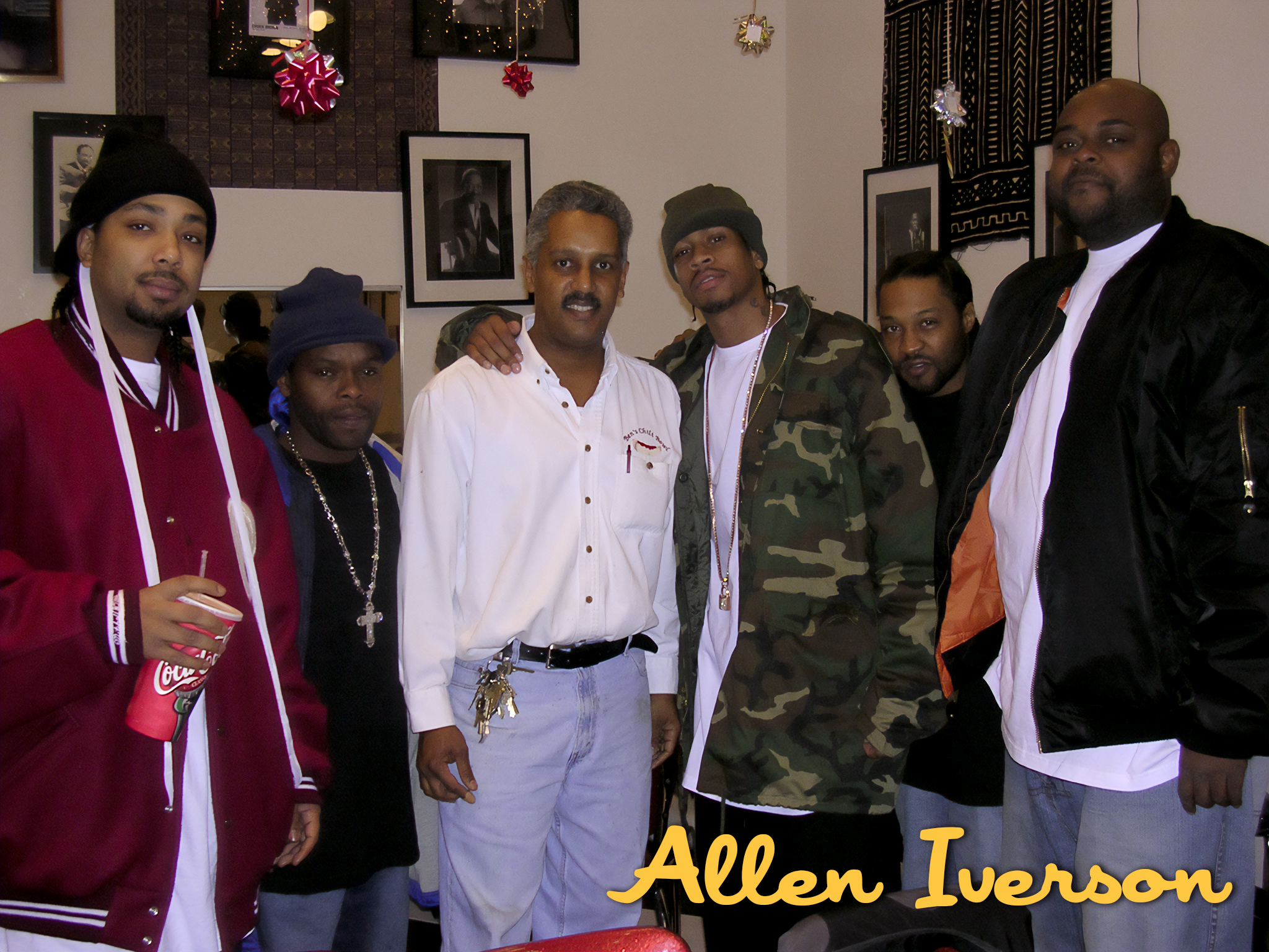 A group of seven men standing indoors, some wearing casual clothing like jackets, camouflage, and accessories, with framed pictures and decorations on the wall behind them, and the text 'Allen Iverson' in yellow at the bottom.