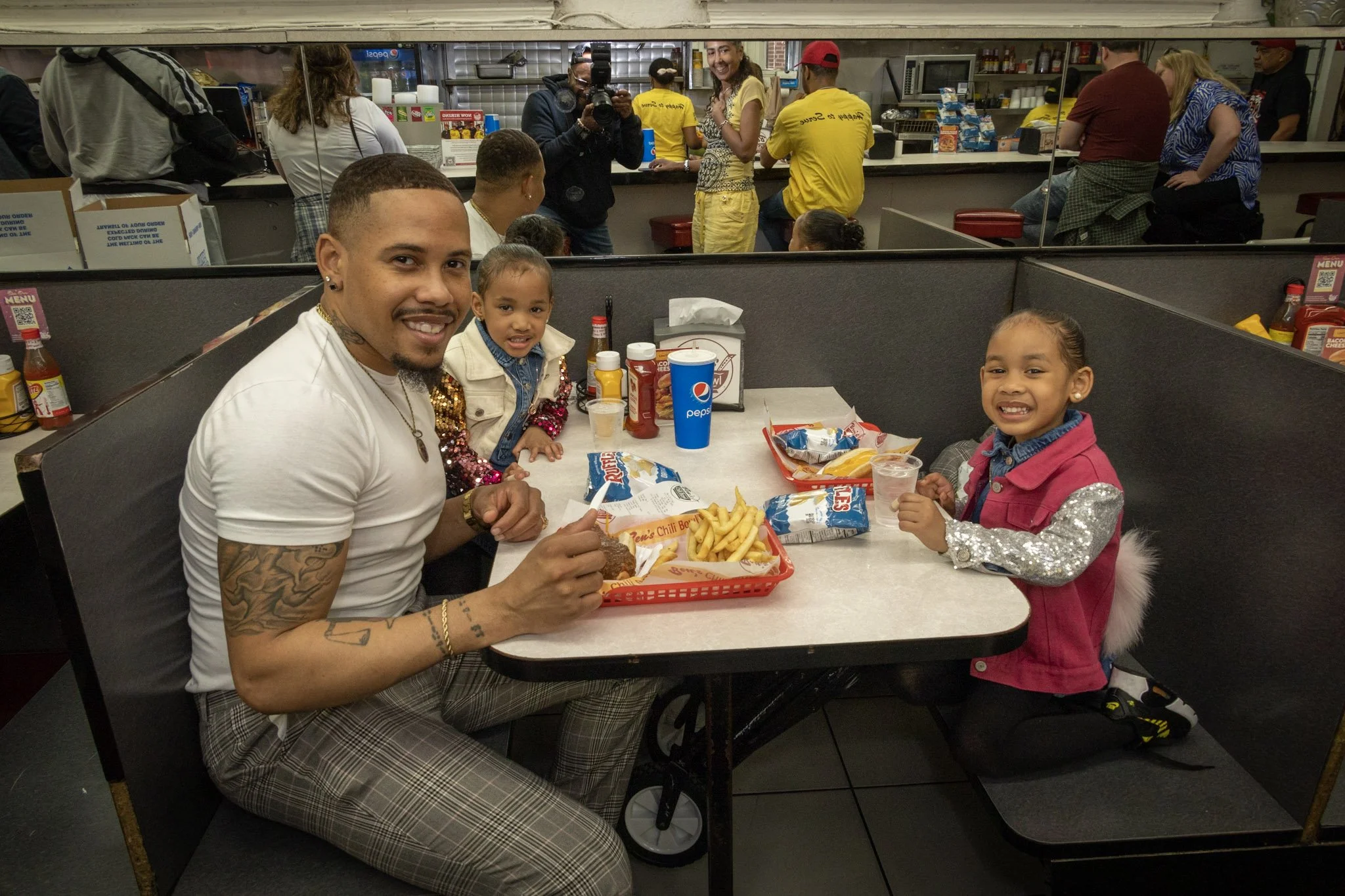 A man with tattoos smiling at the camera sitting with two young girls at a booth in a fast food restaurant, with trays of French fries, burgers, condiments, and a Pepsi in front of them. Other customers and staff are visible in the background.