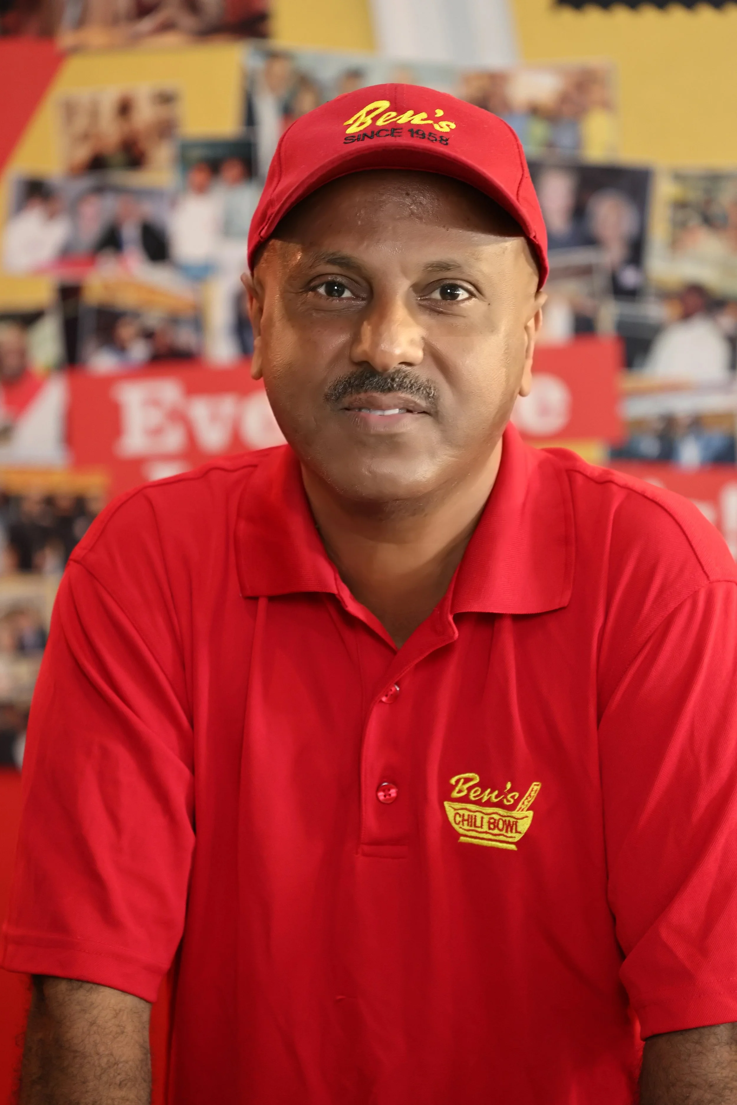 A man wearing a red polo shirt and matching red cap with yellow embroidery, in front of a collage of photos and a red sign that says "Eve". The polo shirt and cap both have the logo "Ben's Chilli Bowl."