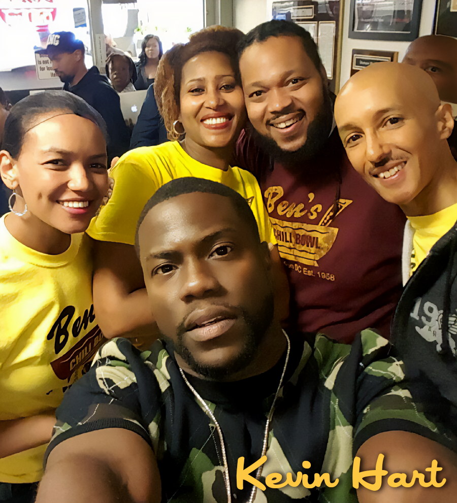 Group of six smiling people taking a selfie in a restaurant or cafe, some wearing yellow and one with a maroon shirt, with a busy background including other patrons and framed pictures on the wall.