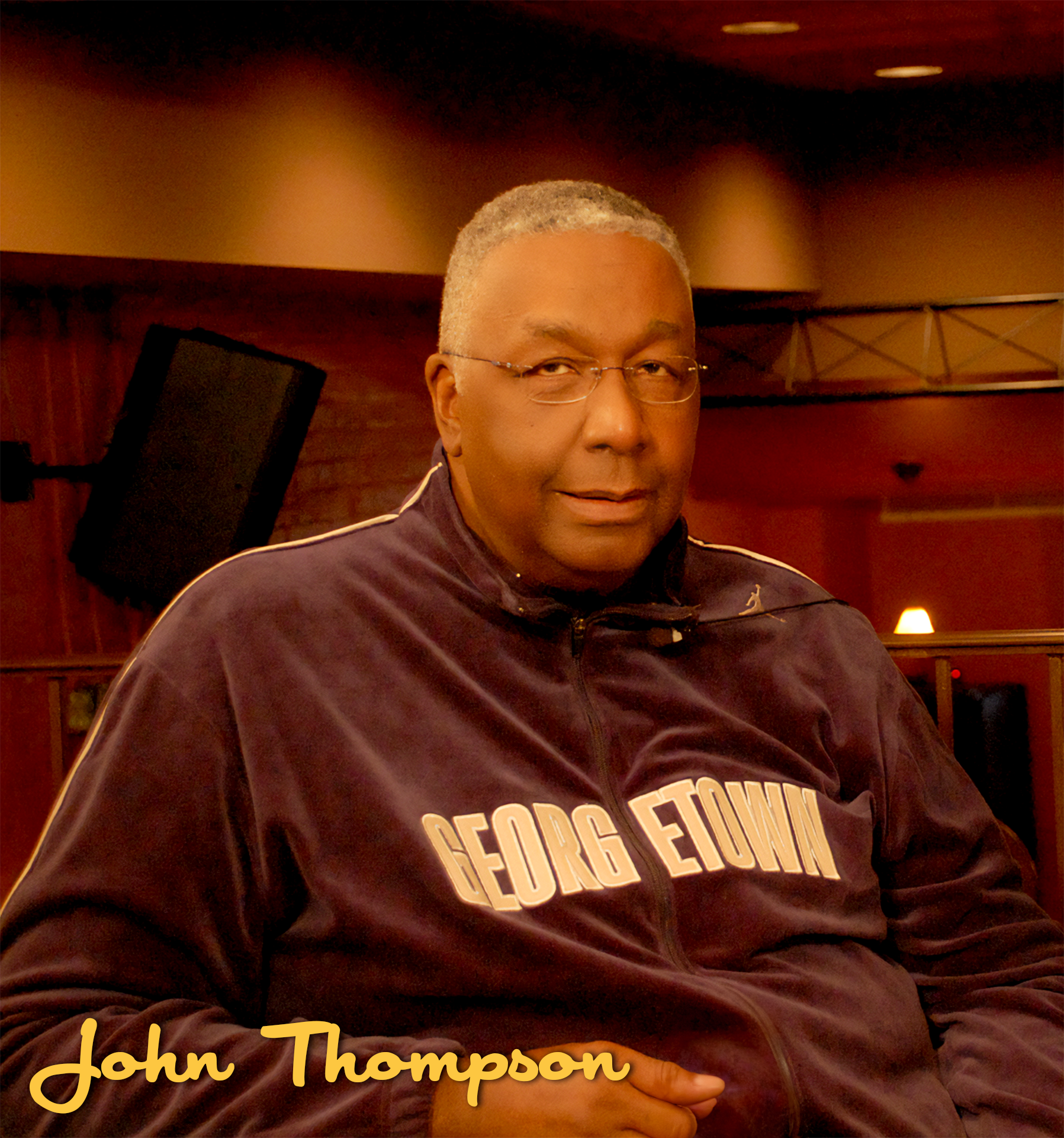 A man wearing glasses and a maroon Georgia State sweatshirt, sitting indoors with a wooden wall and speaker in the background.