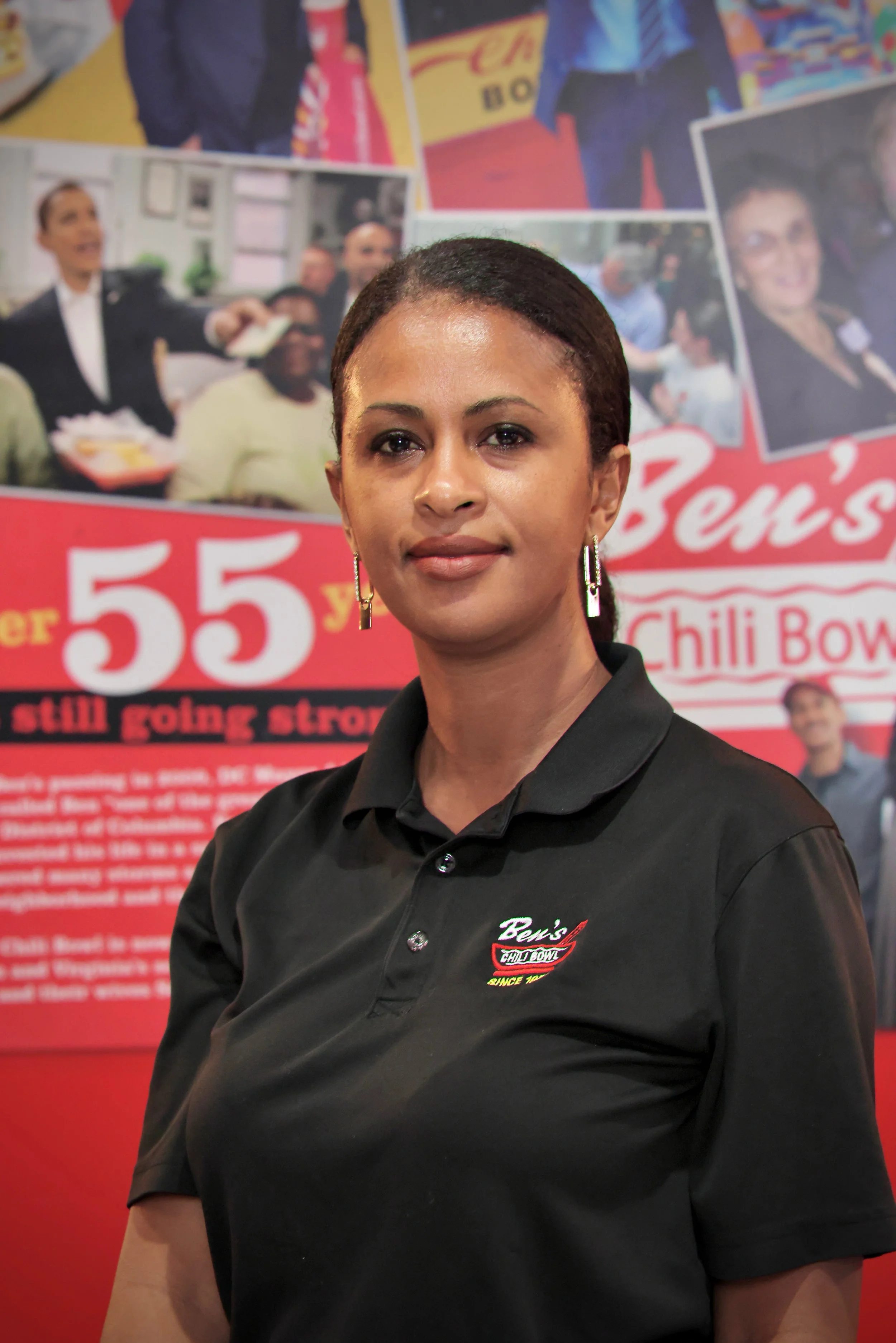 A woman with short black hair, wearing a black collared shirt with a logo that reads Ben's Chili Bowl, stands in front of a red background with photos and text related to Ben's Chili Bowl.