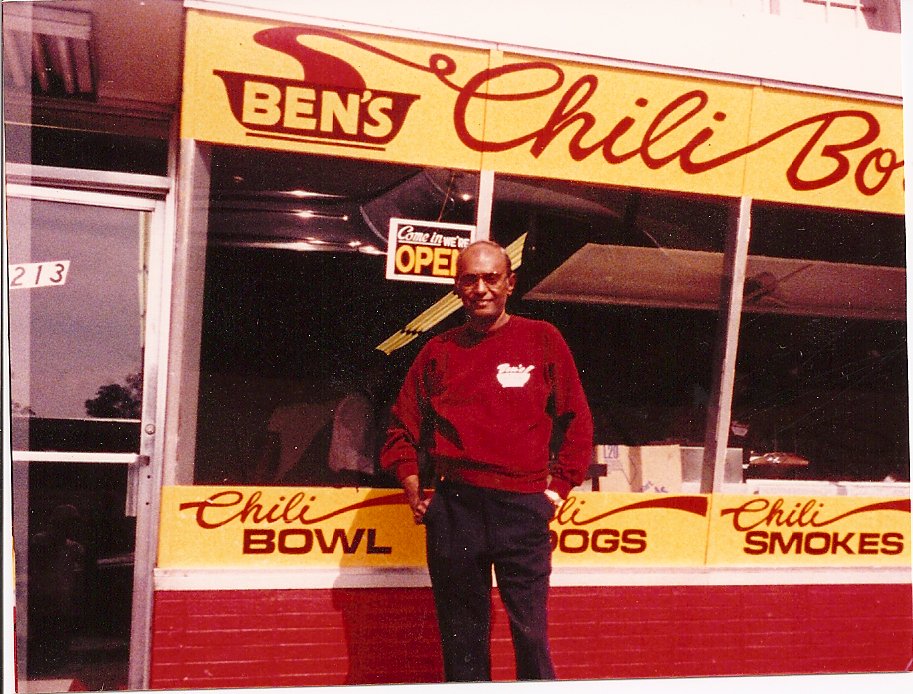 A man standing outside a restaurant named Ben's Chili Bowl, which serves chili bowls, hot dogs, and smoked items. The restaurant has a bright yellow and red sign with a chili pepper graphic and an 'OPEN' sign in the window.