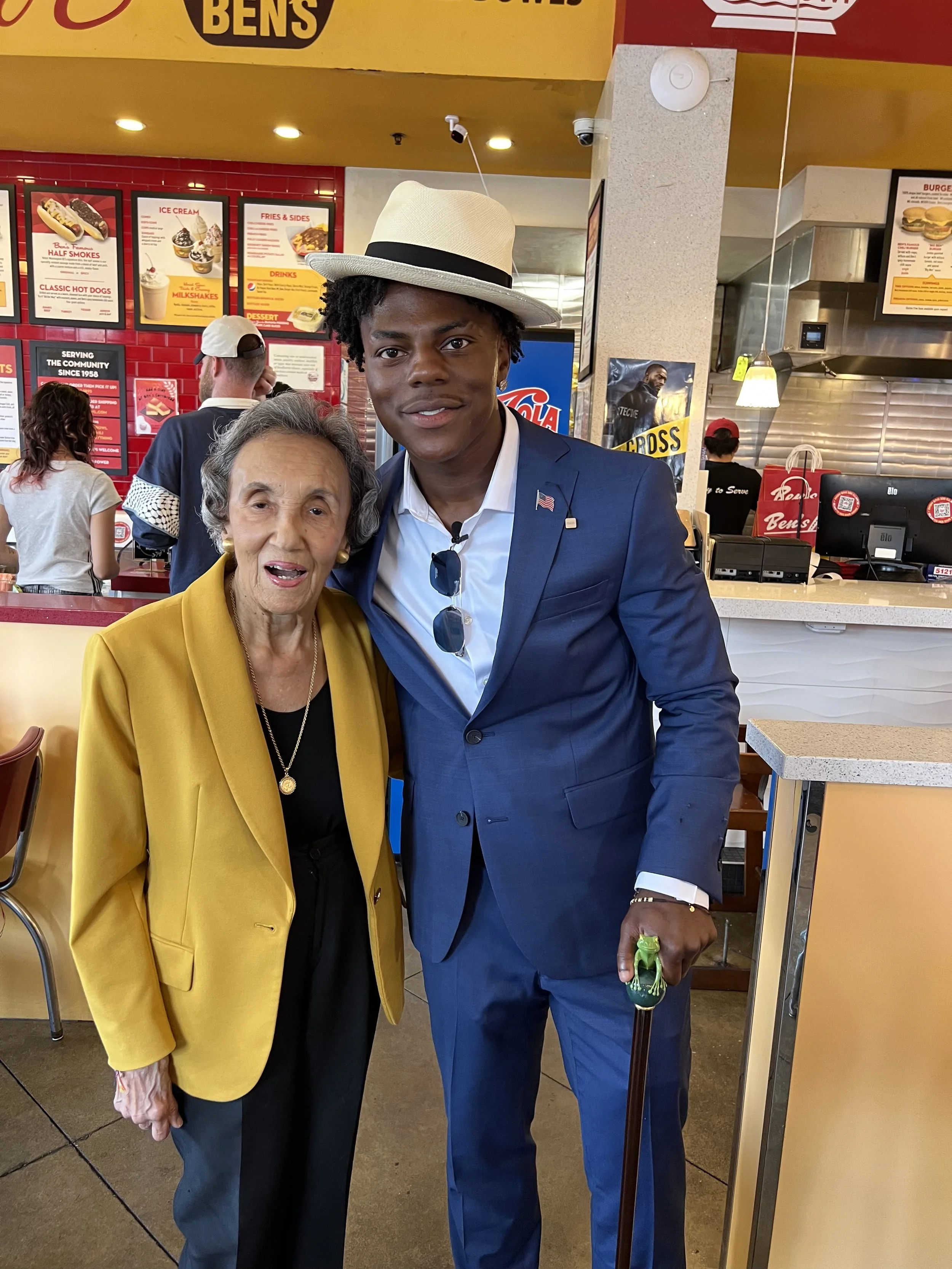 An elderly woman in a yellow blazer standing next to a young man in a blue suit with a cane, inside a fast-food restaurant with a red and yellow menu board in the background.